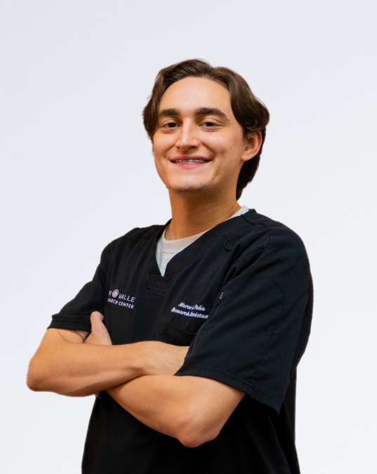 Young man in a black medical scrub top crossing his arms and smiling against a plain white background.
