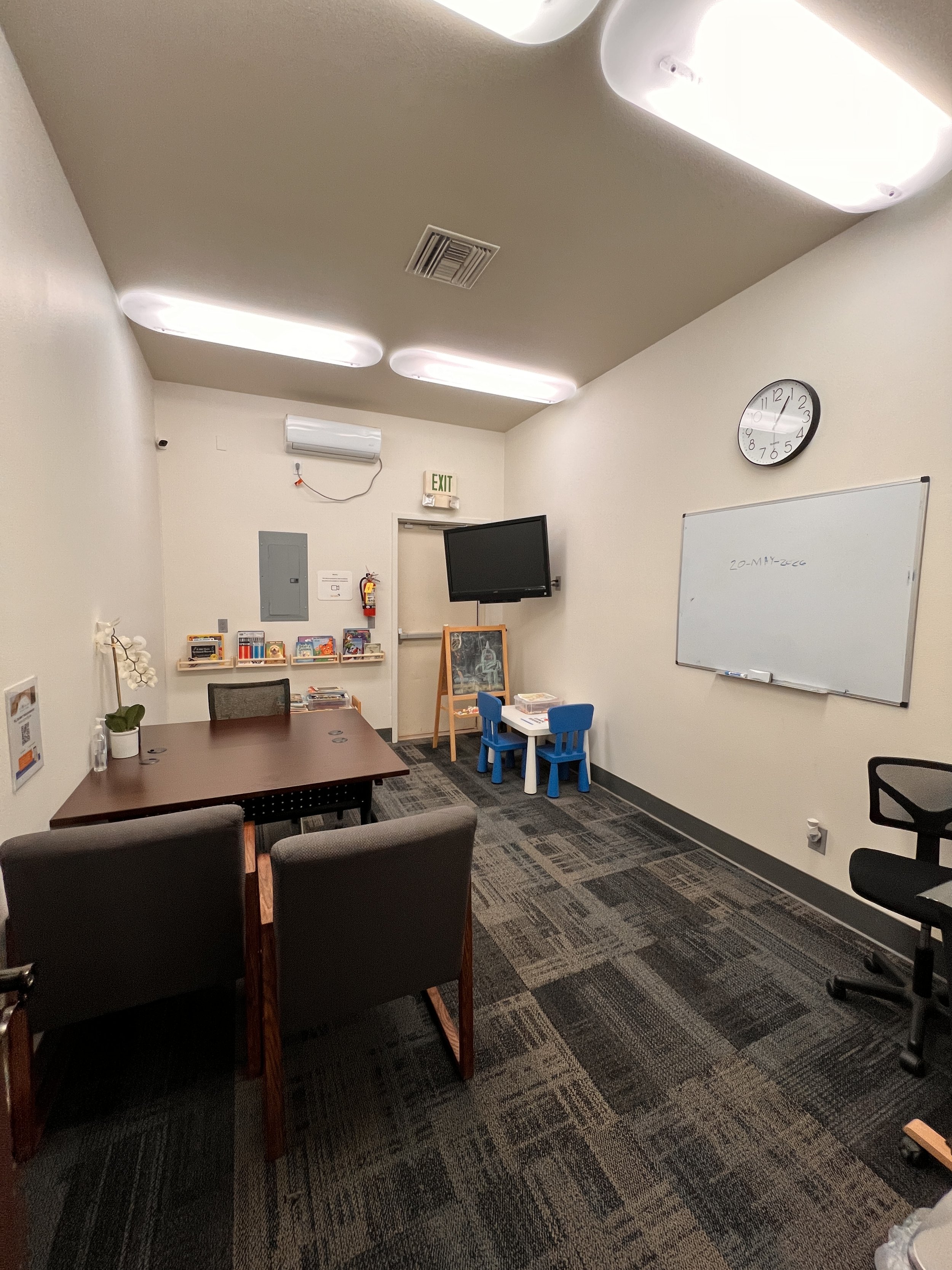 An office or classroom with a whiteboard, a wall clock, a television, a small child-sized table with chairs, bookshelves, and various office supplies. The ceiling has several fluorescent light fixtures.