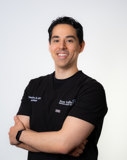Portrait of a smiling man with dark hair, wearing a black medical uniform with Sun Valley logo, standing with arms crossed against a plain white background.