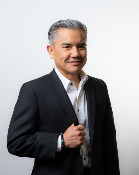 A middle-aged man in a black suit and white shirt standing against a plain white background.