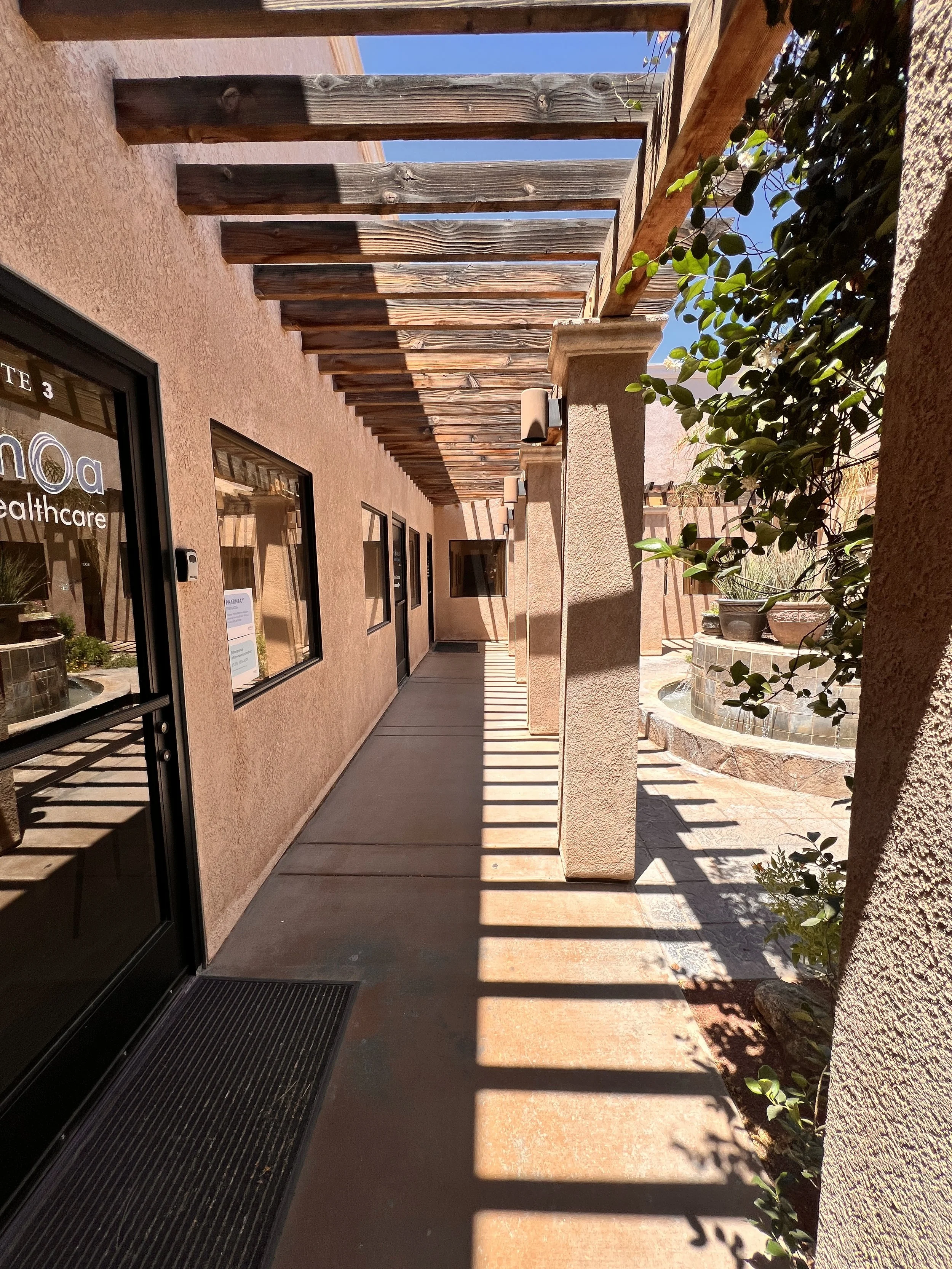 Outdoor walkway with beige stucco walls, wooden pergola casting shadows, windows, plants, and a water fountain visible in the background.