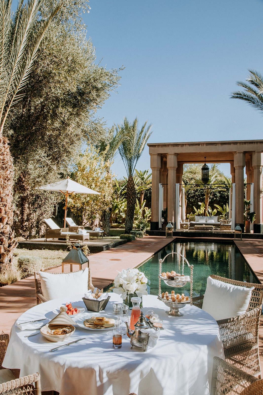 A luxurious outdoor poolside dining area with a round table covered in a white tablecloth, set with plates of food and drinks, surrounded by wicker chairs with white cushions. In the background, there are lounge chairs with umbrellas, lush tropical trees, and a classical-style building with columns and a fountain, all under a clear blue sky.