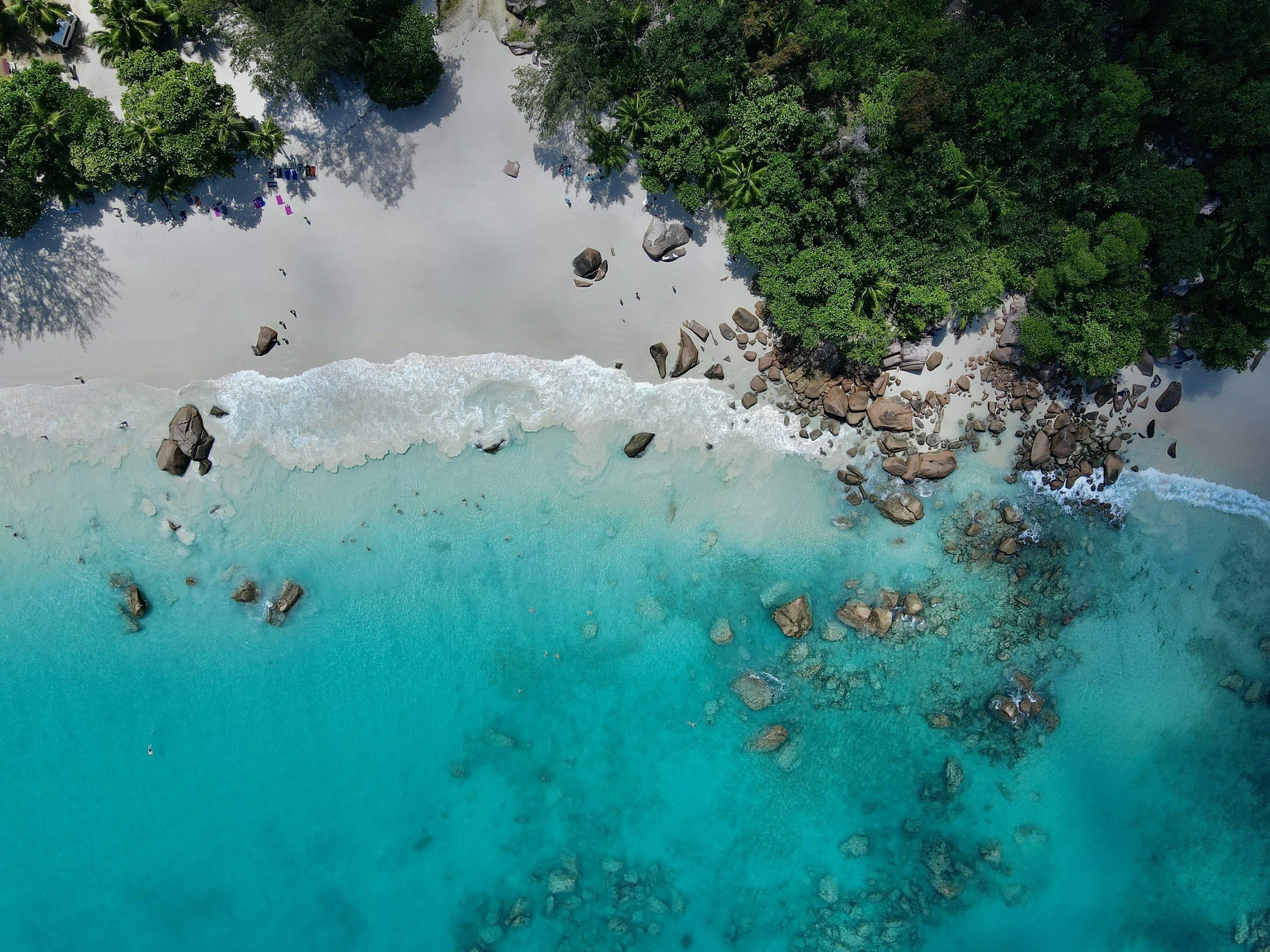 Aerial view of a tropical beach with white sand, turquoise water, large rocks near the shoreline, lush green trees, and some people relaxing in the shade and on the beach. Seychelles.