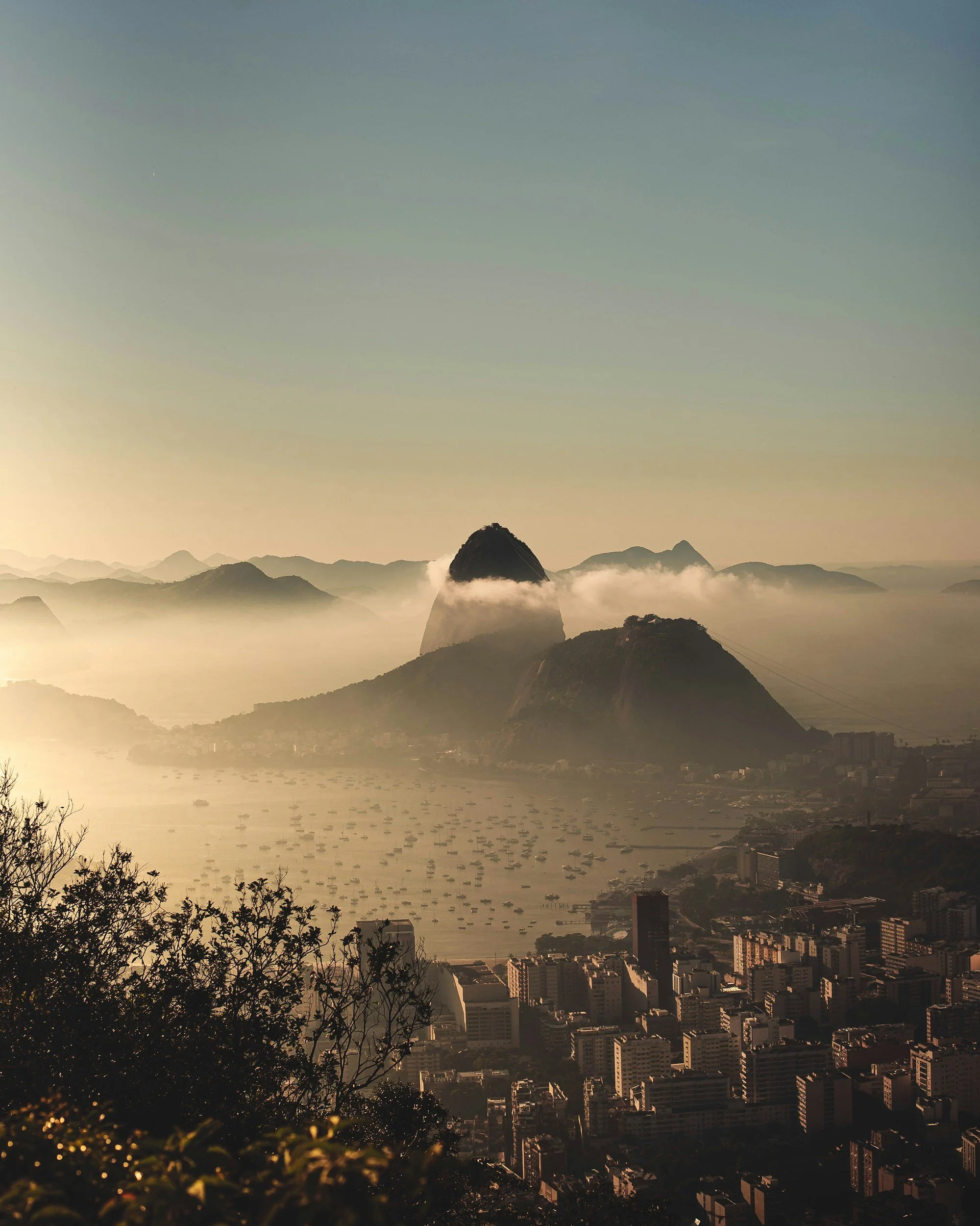View of a city skyline with high-rise buildings, hills, and mountains in the background, with fog or mist covering parts of the landscape, possibly at sunrise or sunset. Rio de Janeiro. Brazil