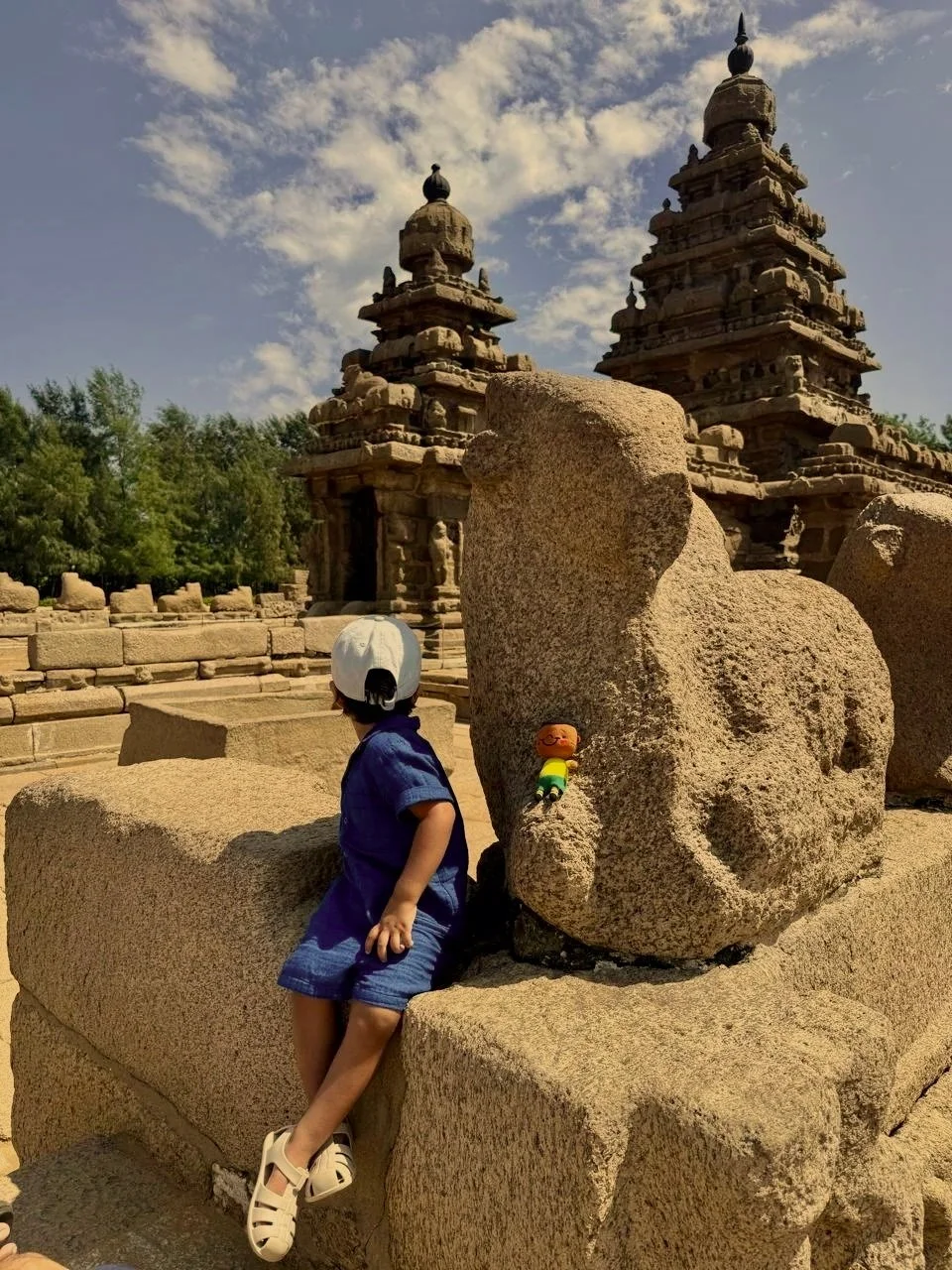 A young boy in a blue shirt and shorts sitting on large sandstone blocks at an ancient temple site with stupa-like structures and a partly cloudy sky in the background. India