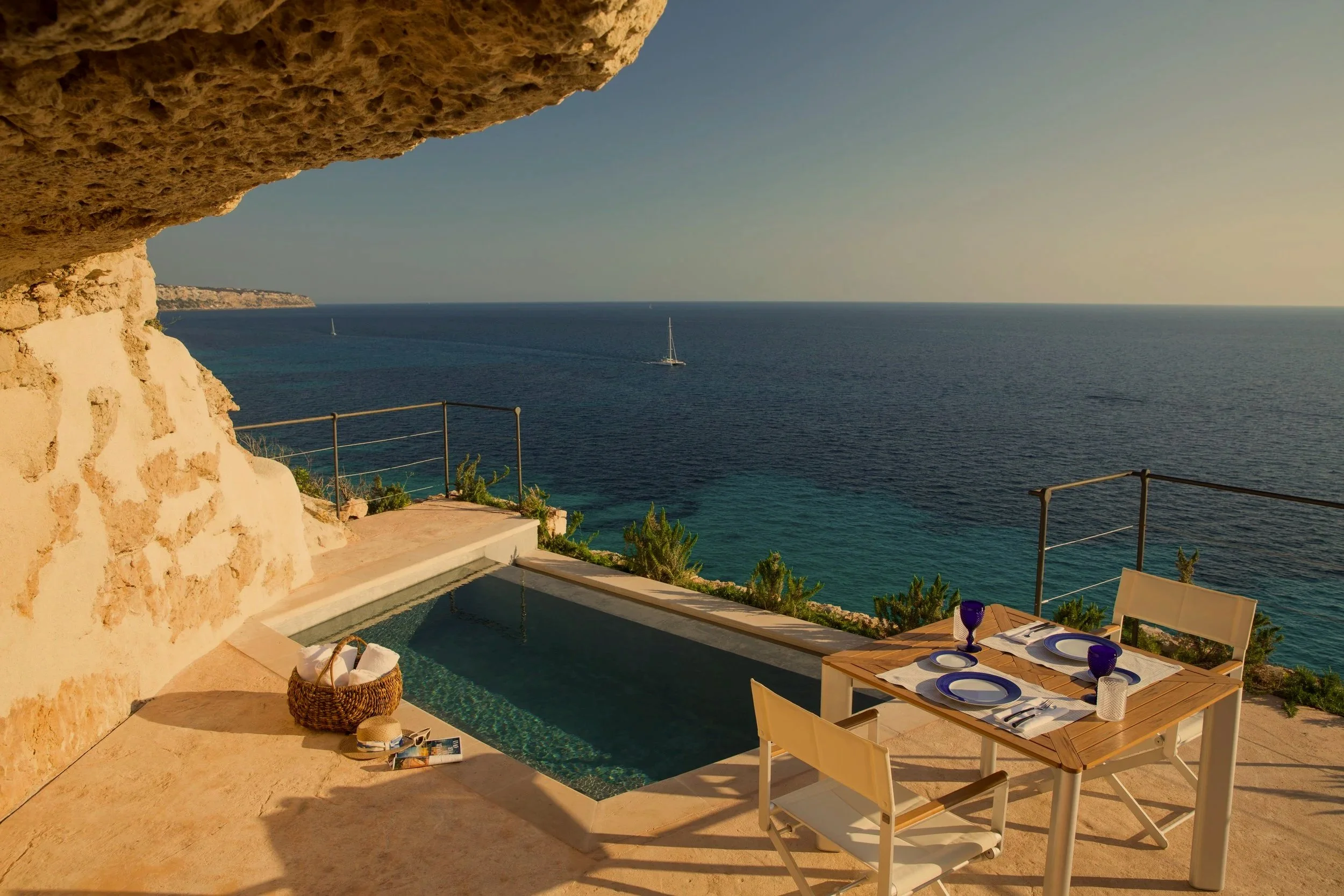 Outdoor dining area with a table set for four, overlooking the ocean, with sailboats in the distance, a stone wall on the left, and a swimming pool in the foreground. Mallorca