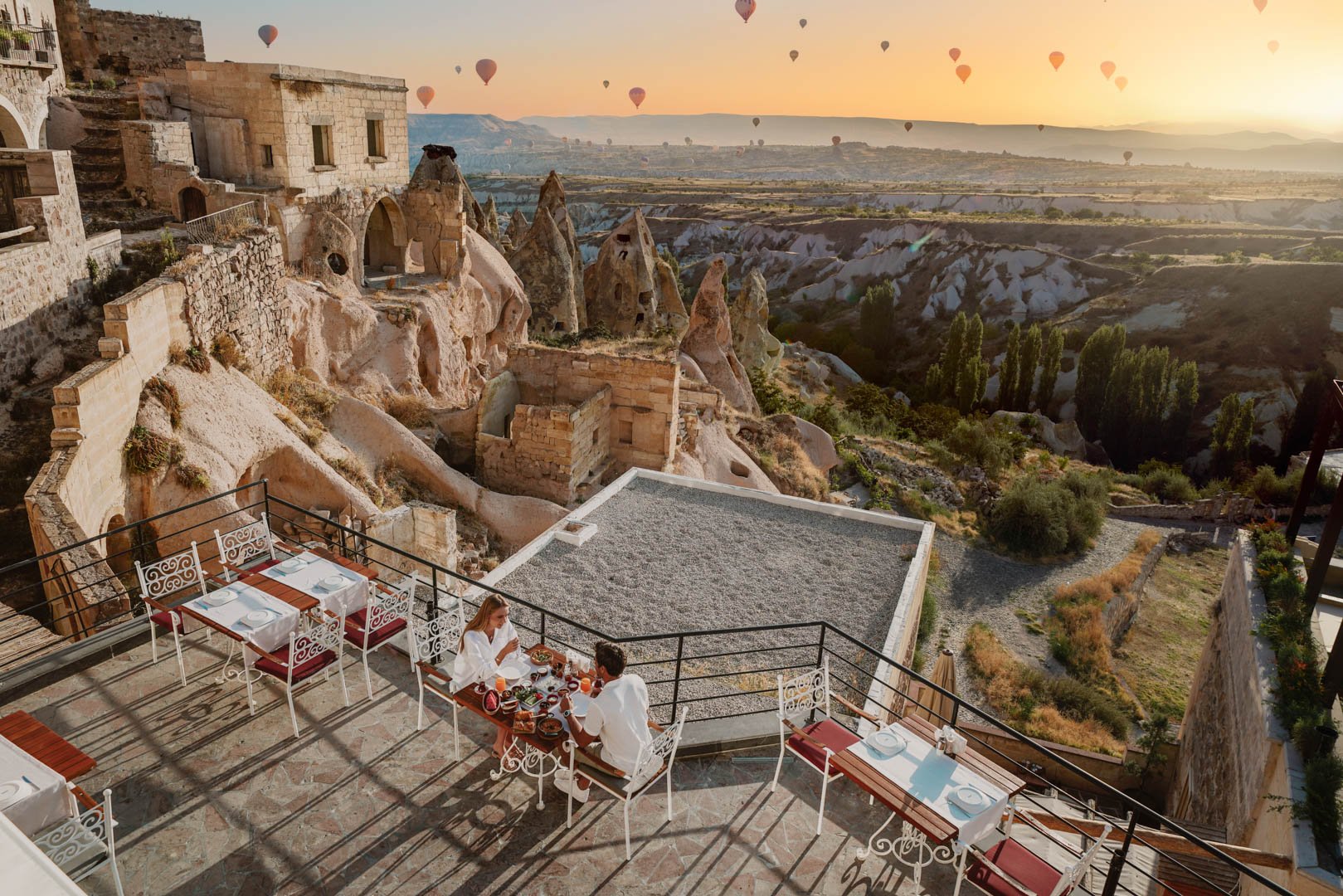 A rooftop restaurant with a table for two set for dining, overlooking a landscape of ancient rock formations and several hot air balloons in the sky at sunset in Cappadocia, Turkey.