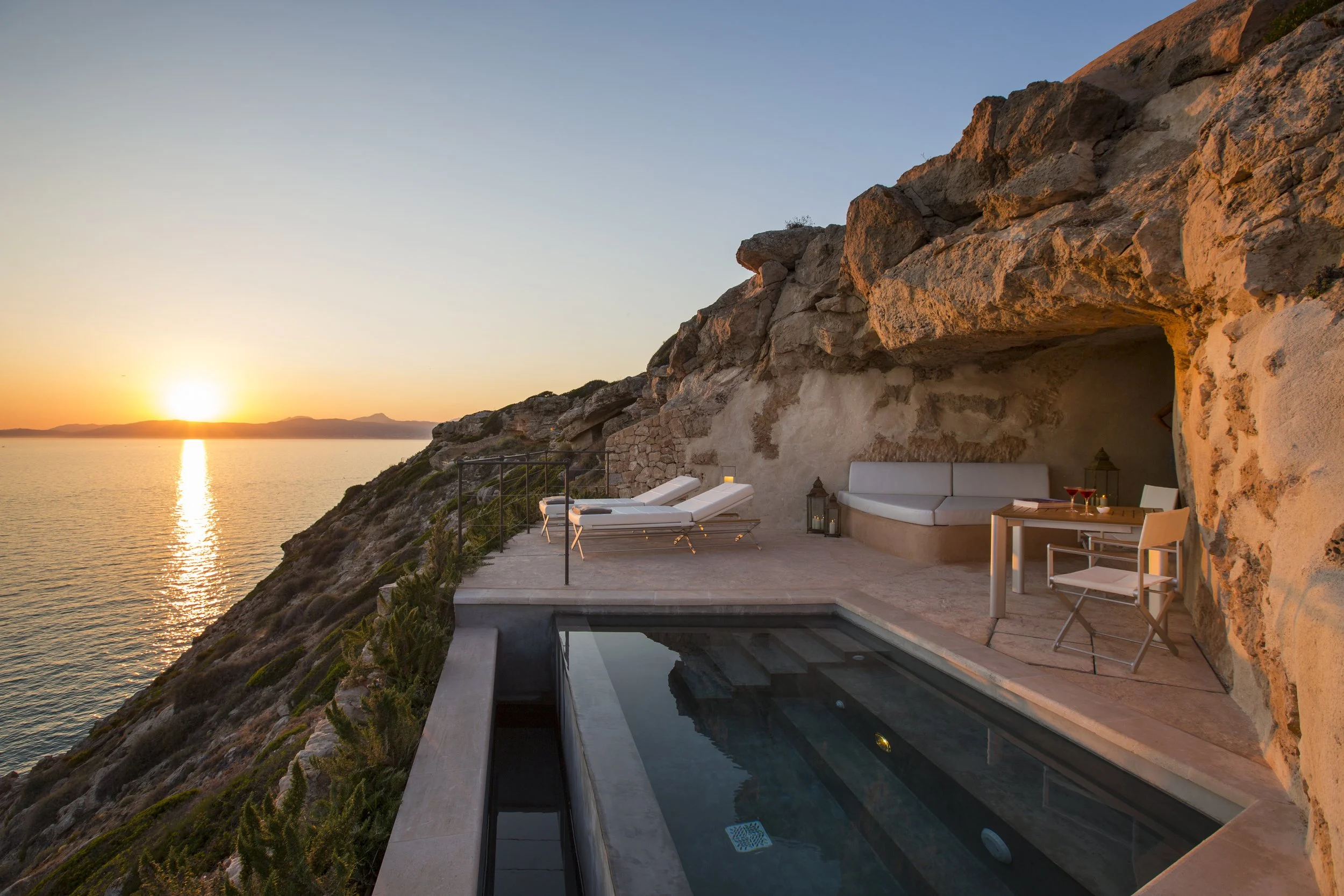 A terrace overlooking the ocean at sunset, with lounge chairs, a small pool, and seating area built into a rocky cliff. Mallorca. Cap Rocat