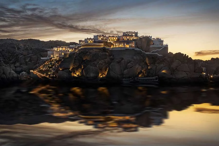 A large, luxurious hotel or resort built on a rocky hill at dusk, with outdoor lighting illuminating the structure and its surroundings, reflecting in the water below. BenBen. Egypt