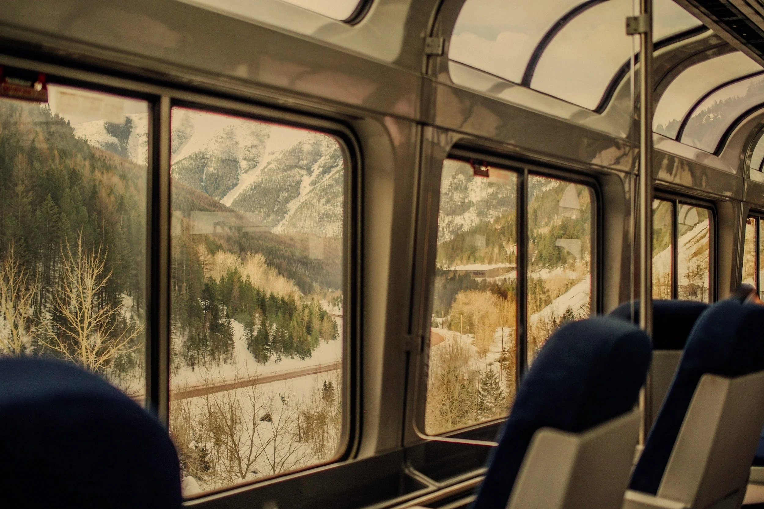 Inside a train, looking out the window at snow-covered mountains and trees during daytime.