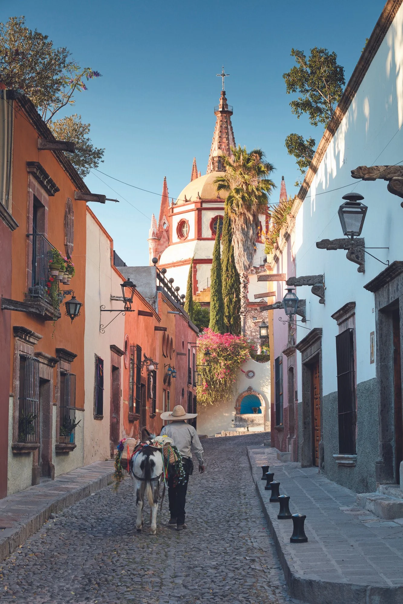 A cobblestone street in a colorful Mexican town with a person walking a horse, colonial-style buildings with iron balconies, and a church with a tall steeple in the background, under a clear blue sky. Mexico