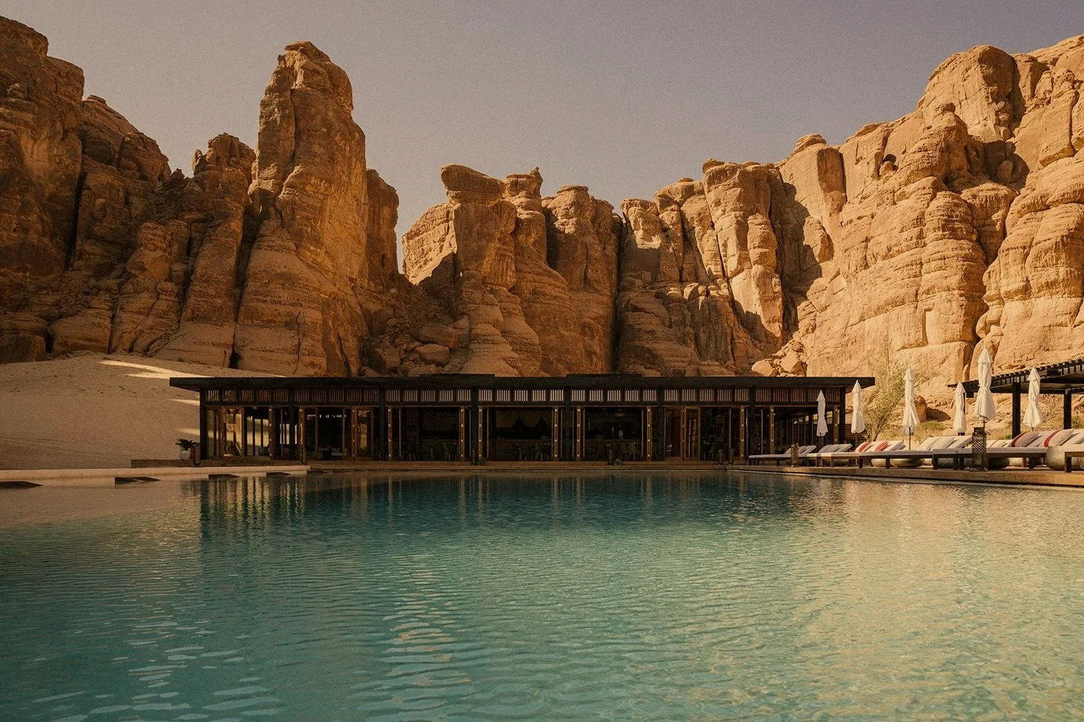 A luxurious infinity pool with lounge chairs and umbrellas, set against a backdrop of towering sandstone cliffs during sunset. Saudi Arabia