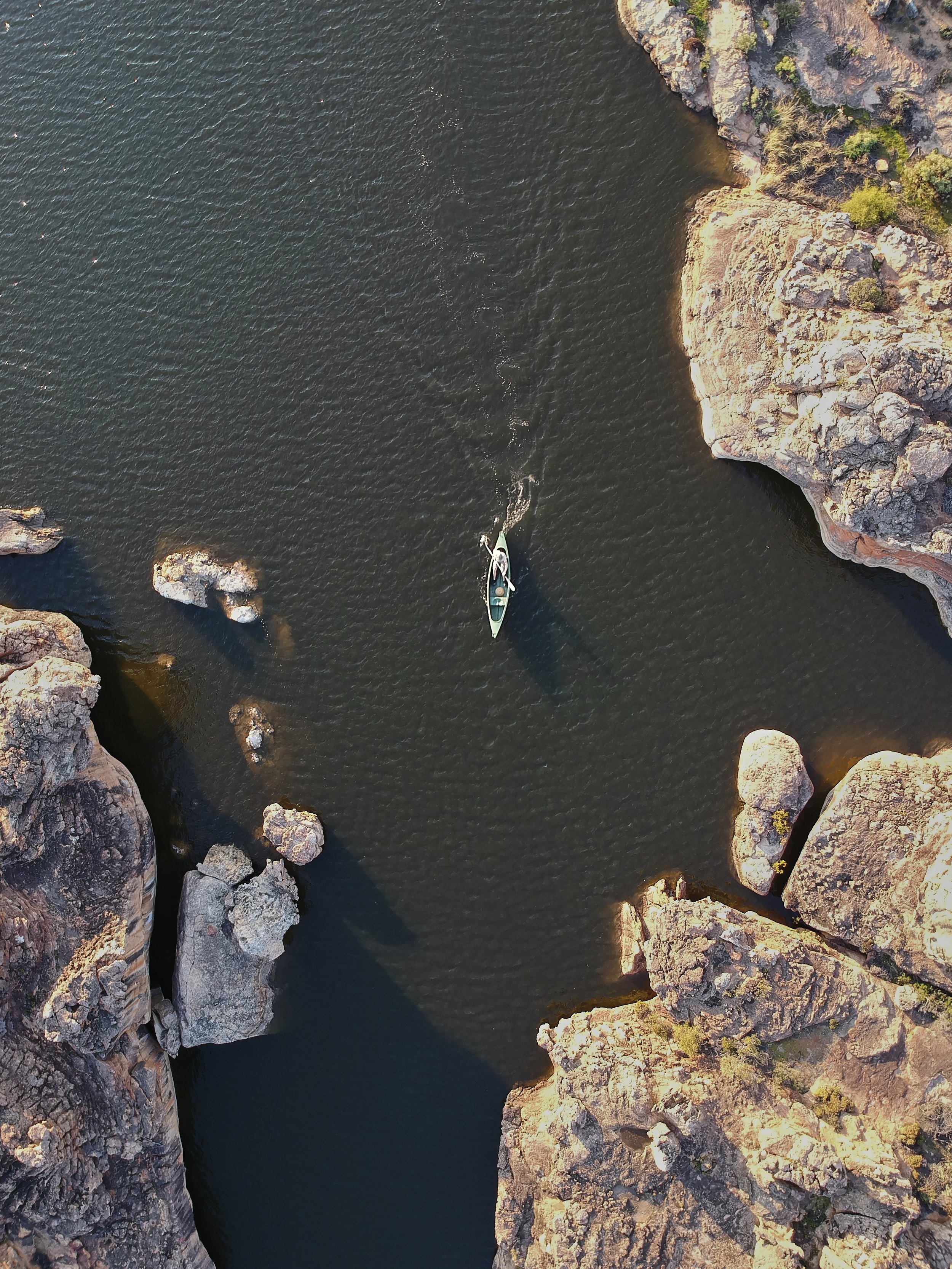 An aerial view of a narrow river or canyon with dark water, surrounded by large rocky cliffs and some sparse vegetation. Africa