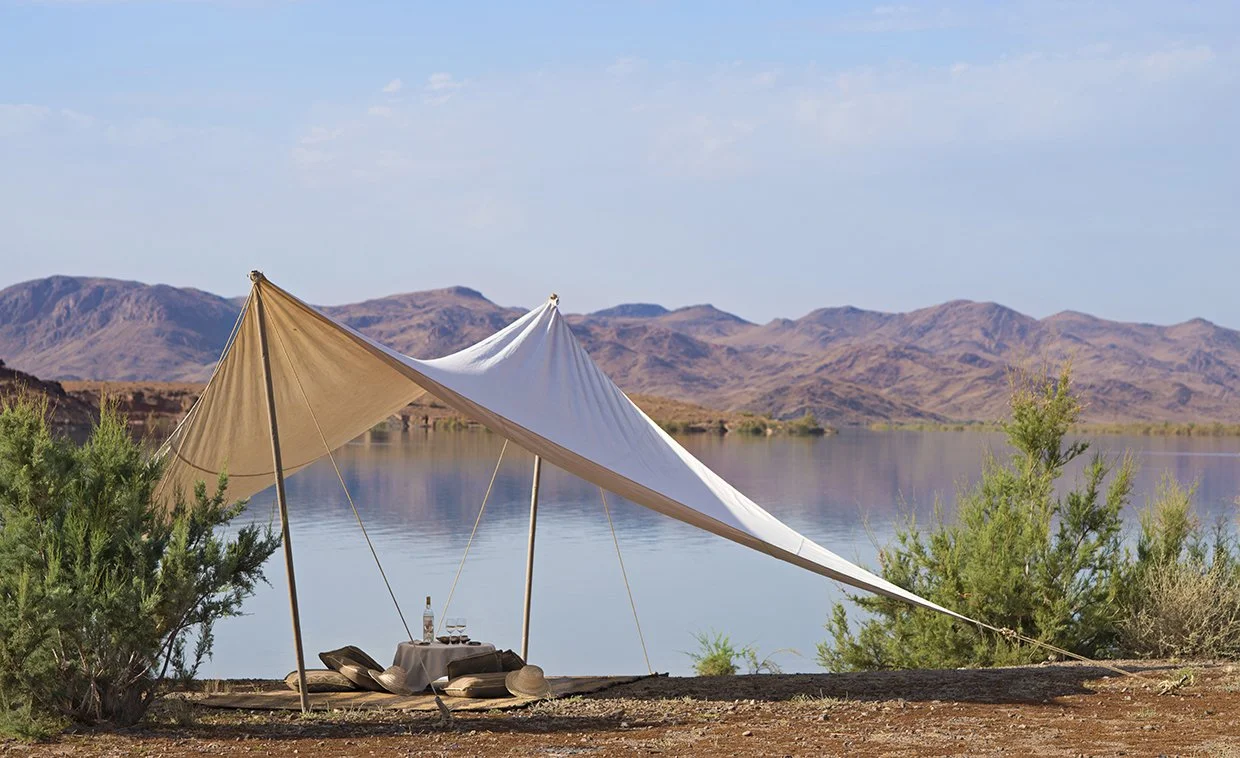 A lakeside scene with a beige and white tent, a picnic setup with a bottle and wine glasses, and mountains in the background. Morocco
