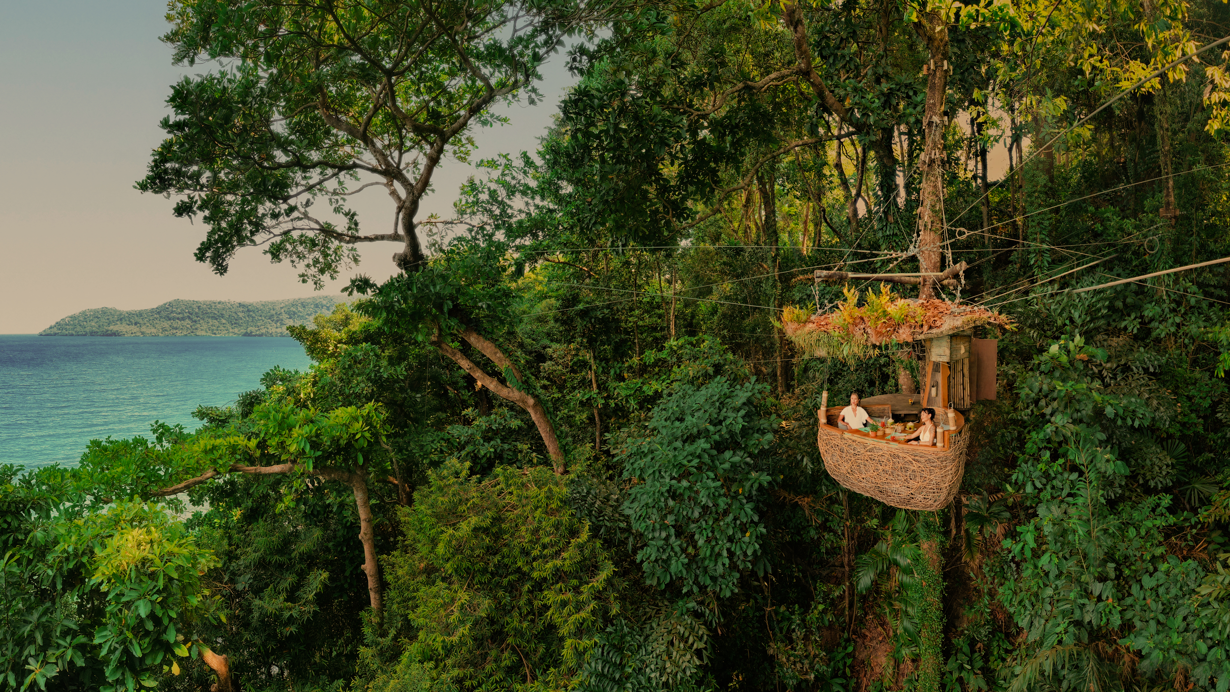 A treehouse with a balcony is built among tall trees in a lush forest near a body of water, with two women sitting at a table on the balcony. Thailand