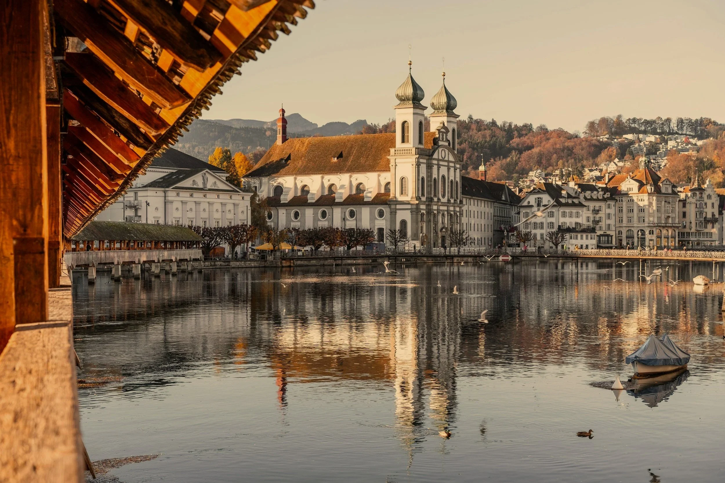 A scenic view of a waterfront town with historic buildings, including a prominent white church with twin onion domes, set against a backdrop of hills and trees with autumn foliage, calm water and a small boat. Lucerne.Switzerland.