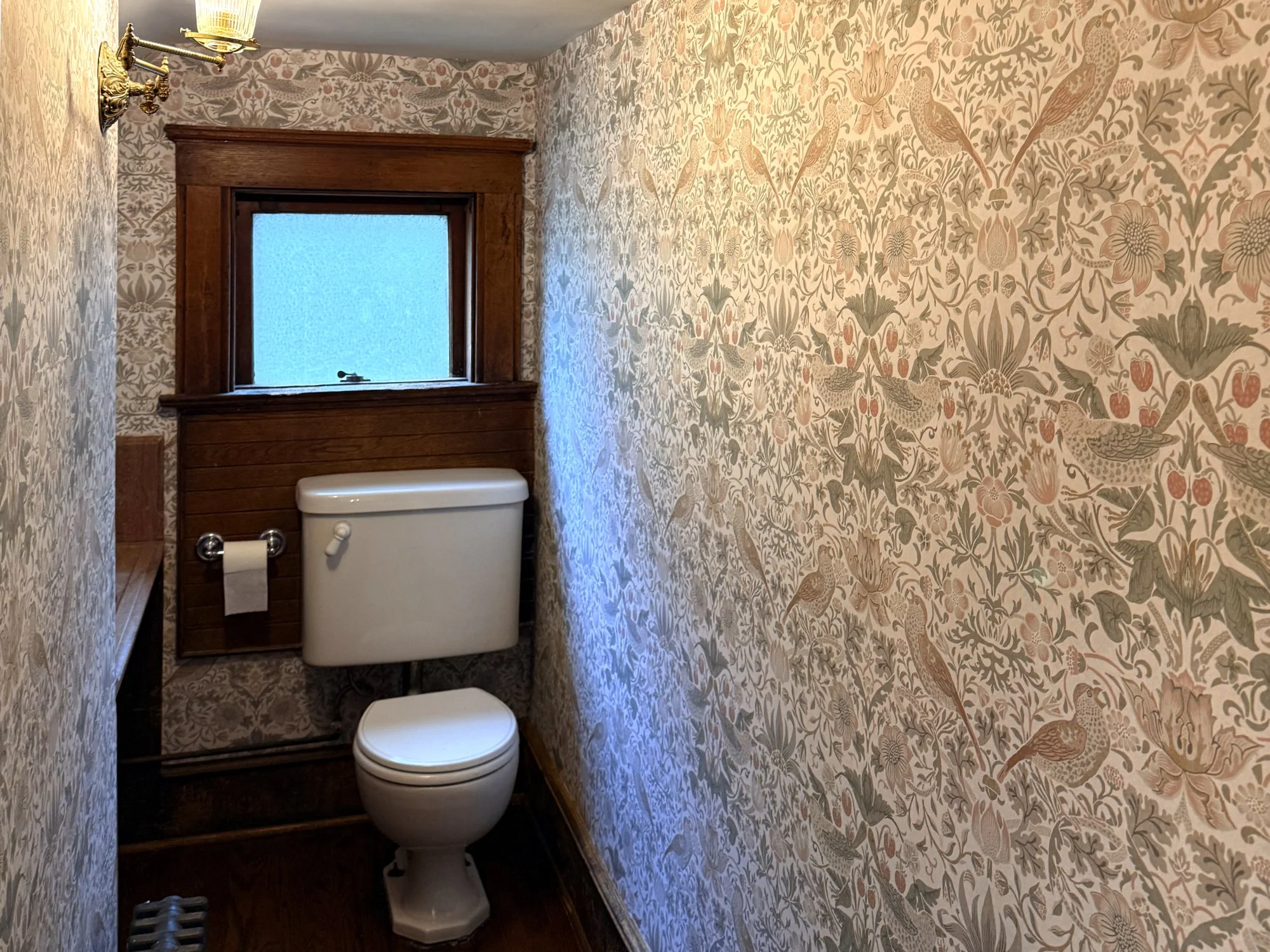 Small vintage bathroom with floral wallpaper, a wood-framed frosted window, a white toilet, and a brass wall sconce light fixture.
