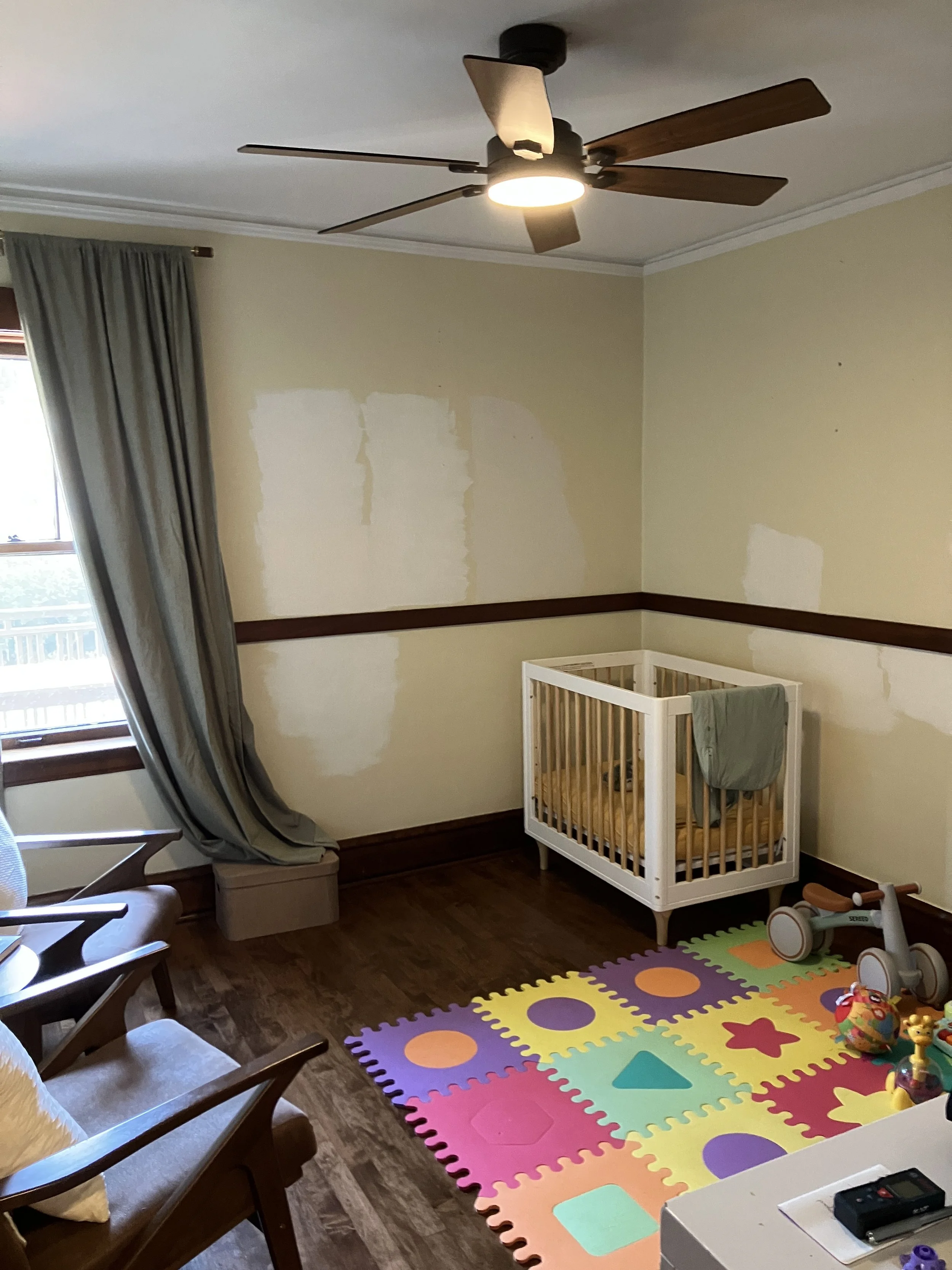 A room with a window and curtain, a wooden floor, a ceiling fan with lights, an unfinished wall with patches of drywall, a colorful foam puzzle mat on the floor, toys including a plastic ball, a ride-on toy, and a crib