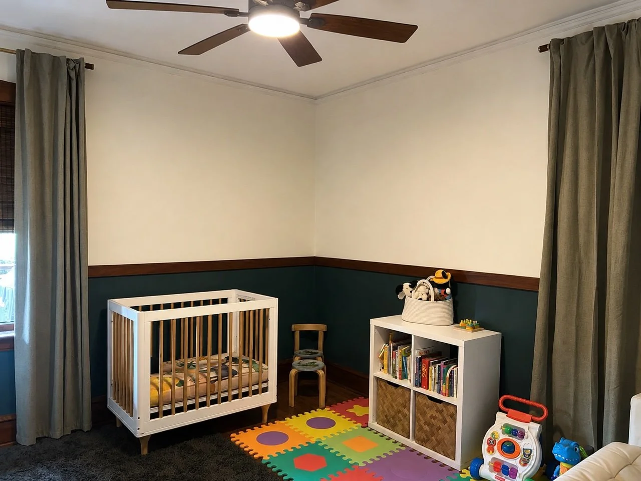 Nursery room with a crib, small chair, bookshelf with books and toys, colorful foam floor mats, and curtains.