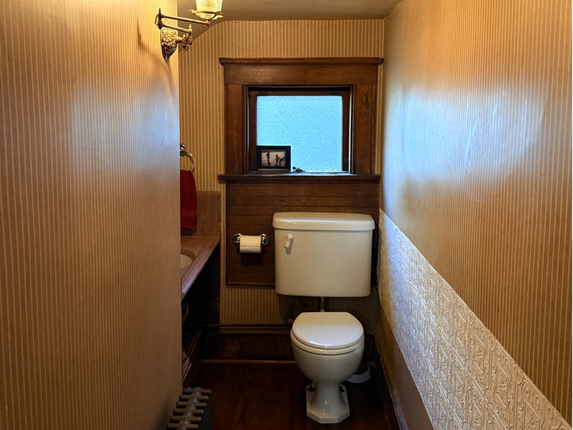 A small bathroom with a toilet, a window with a wooden frame, a framed picture on the window sill, a towel ring with a red towel, and patterned wallpaper.