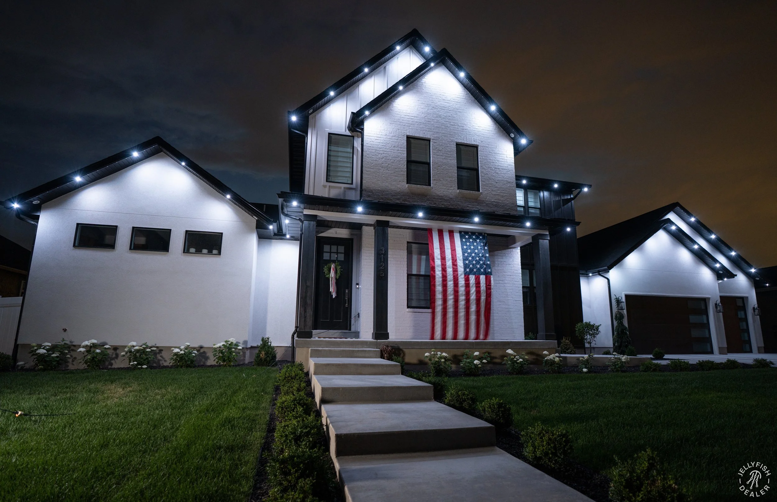 A modern house at night decorated with exterior lighting. The house has white brick and siding, dark window frames, and an American flag hanging from the front porch. The lawn is well-kept with a concrete walkway leading to the front door.