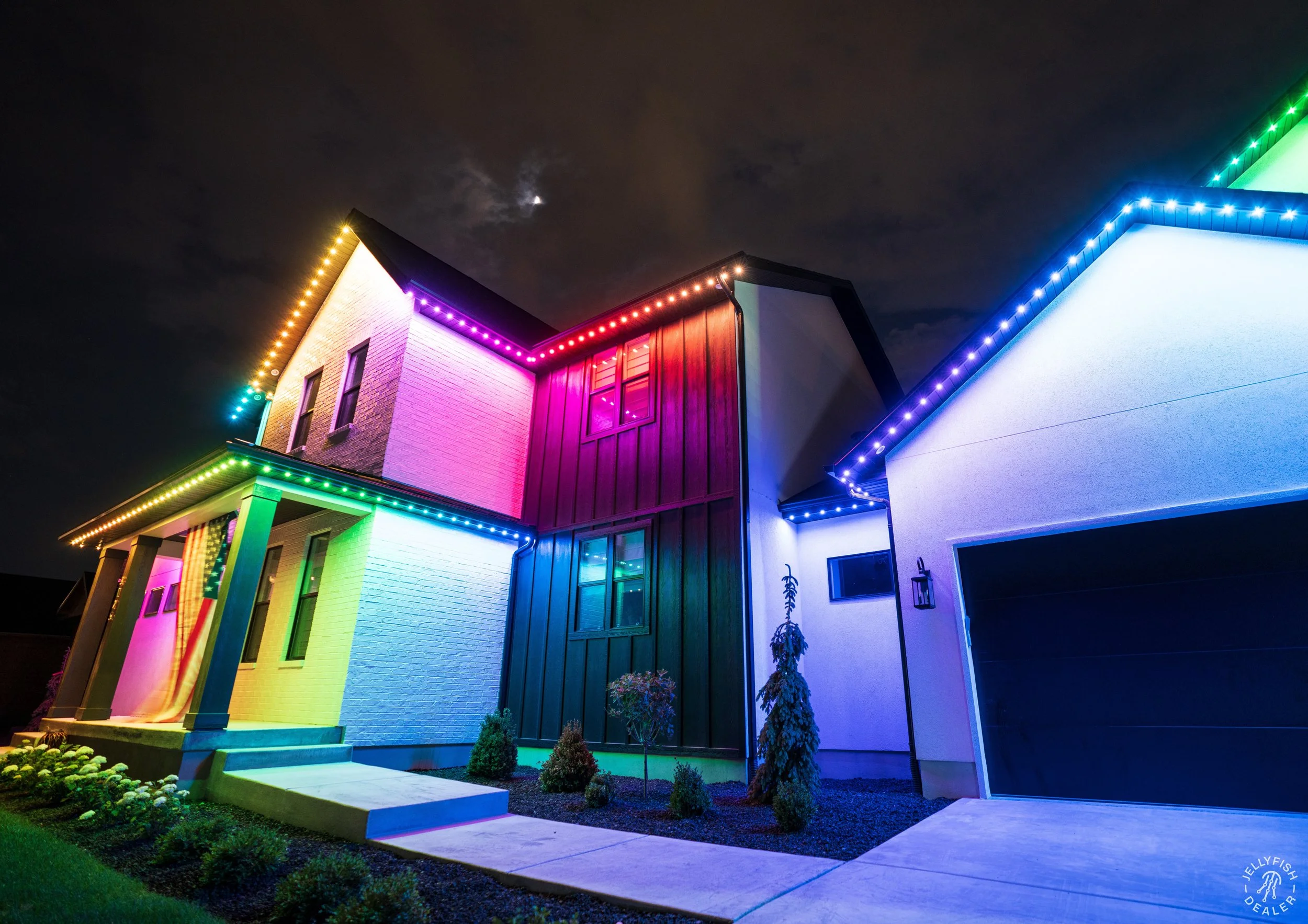 Colorfully lit house at night with rainbow LED lights outlining roof and windows, small plants in front and cloudy sky above.