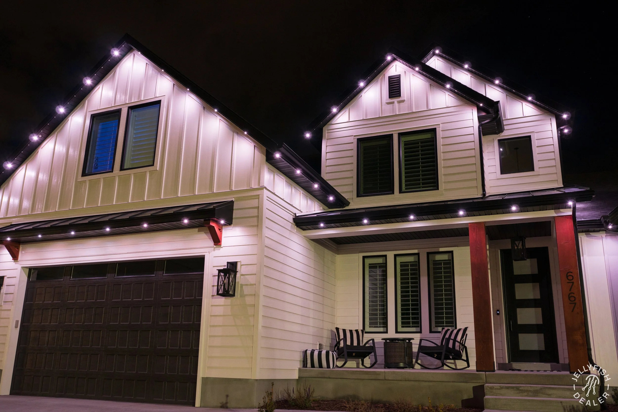 Night view of a modern two-story house illuminated with exterior lighting, featuring a garage door, large windows with shutters, and a patio area with chairs and a table.