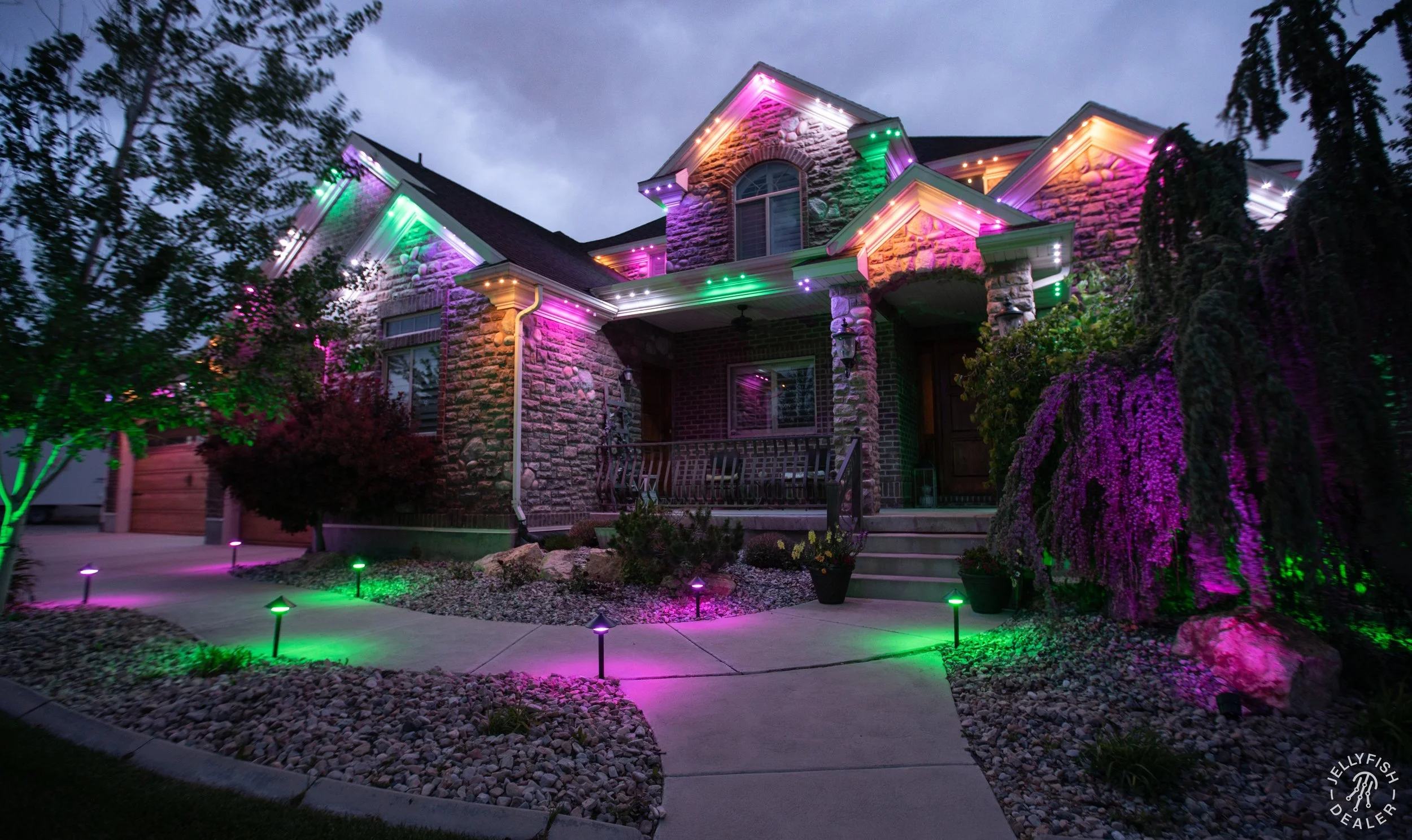 A house decorated with colorful outdoor holiday lights at dusk, with a pathway, landscaped yard, and trees around the house.