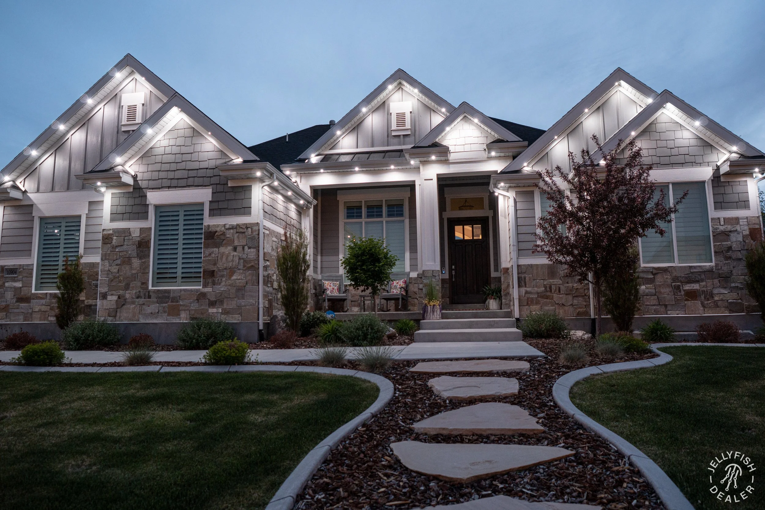 A well-lit modern house at dusk with a landscaped front yard including a curved walkway, small trees, and shrubs.
