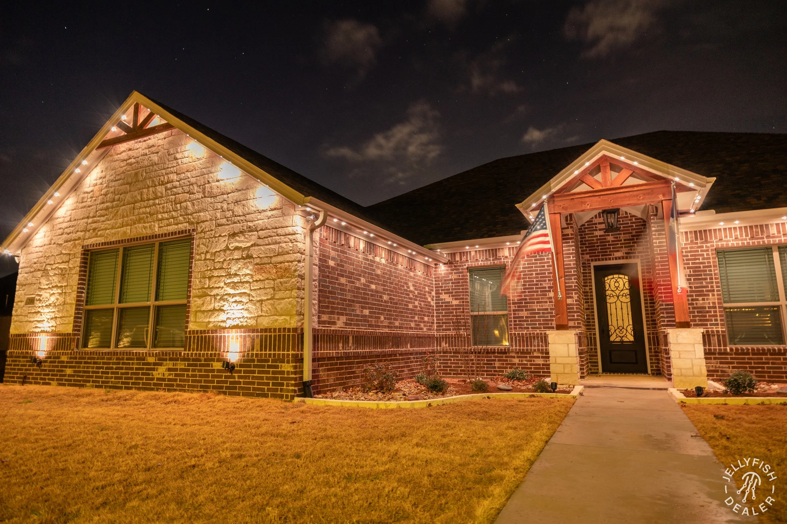 A two-story brick house at night decorated with string lights along the roofline, with American flags near the entrance, a dark shingled roof, and a manicured lawn and walkway in the front yard.