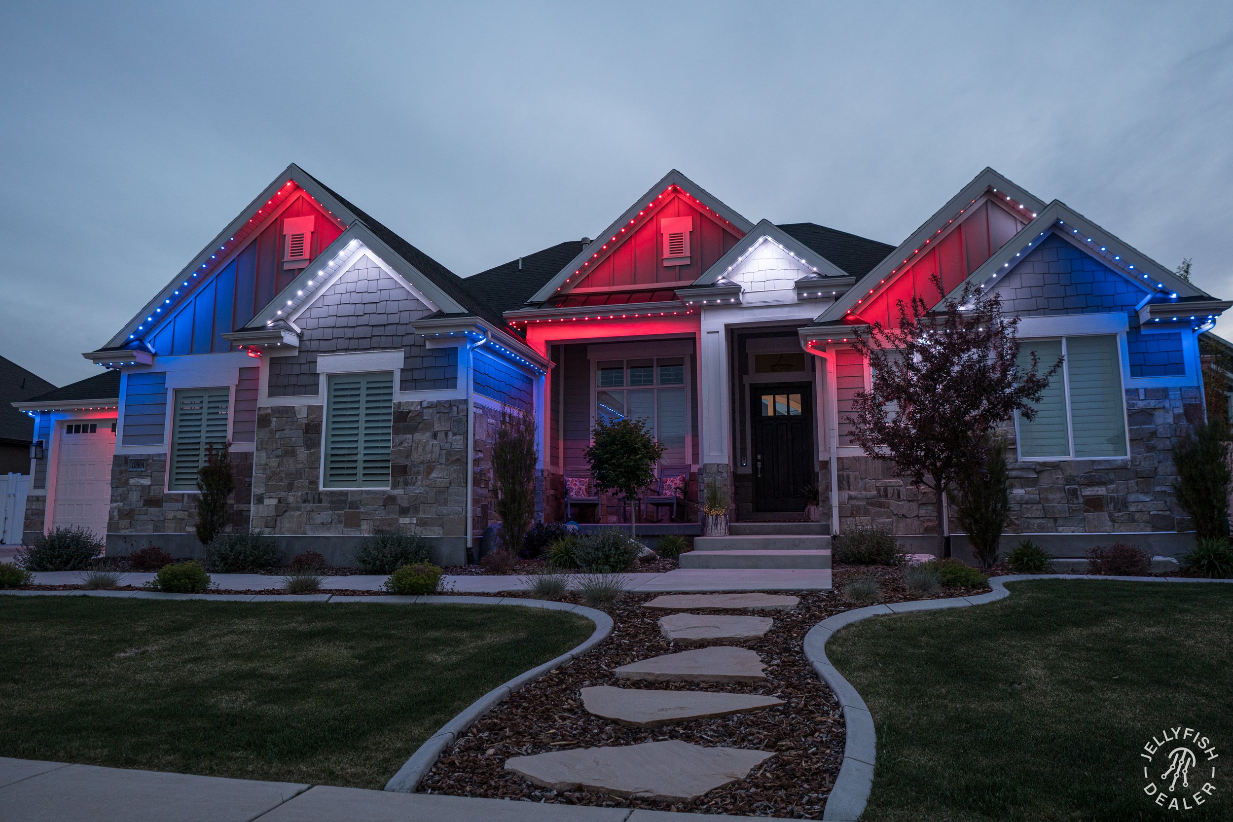 A house decorated with red, white, and blue holiday lights outlining the roof and eaves, with a front yard featuring a winding pathway leading to the front door, and a small tree to the right of the entrance.