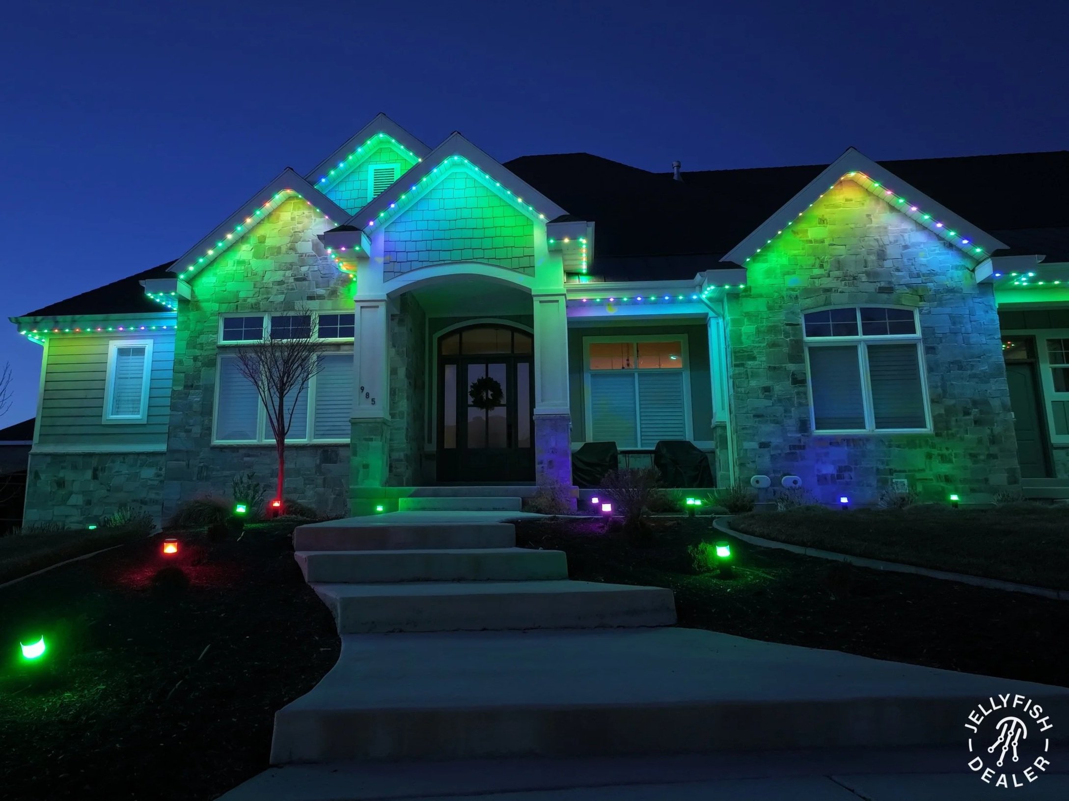 The image shows a house at night decorated with colorful Christmas lights outlining the roof and windows. The house has brick and siding exterior, with a small tree and lit pathway in the front yard.