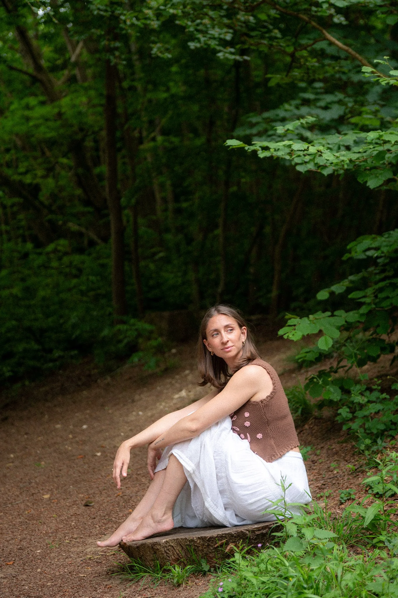 A woman with long hair sitting on a tree stump in a wooded area, wearing a brown tank top and white skirt, looking to her right with a calm expression.