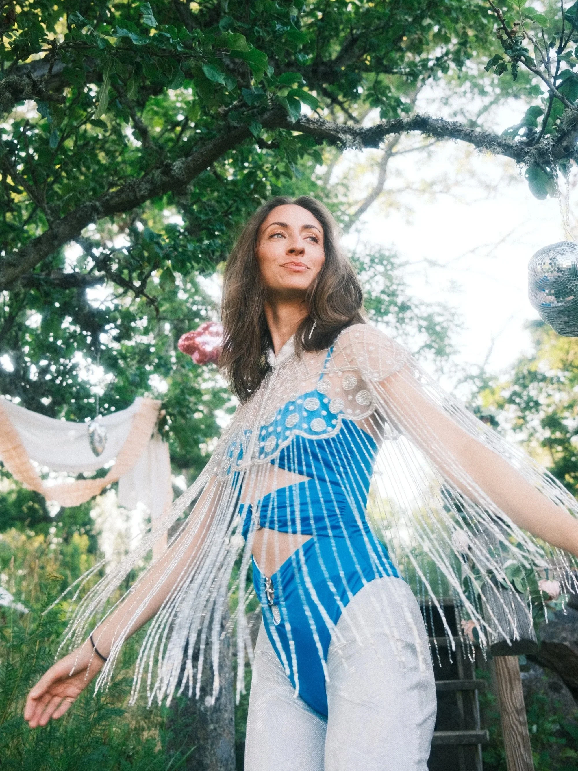 A young woman with dark hair, dressed in a blue and white, stands outdoors under green leafy trees during daytime with her eyes looking upward and to the side.