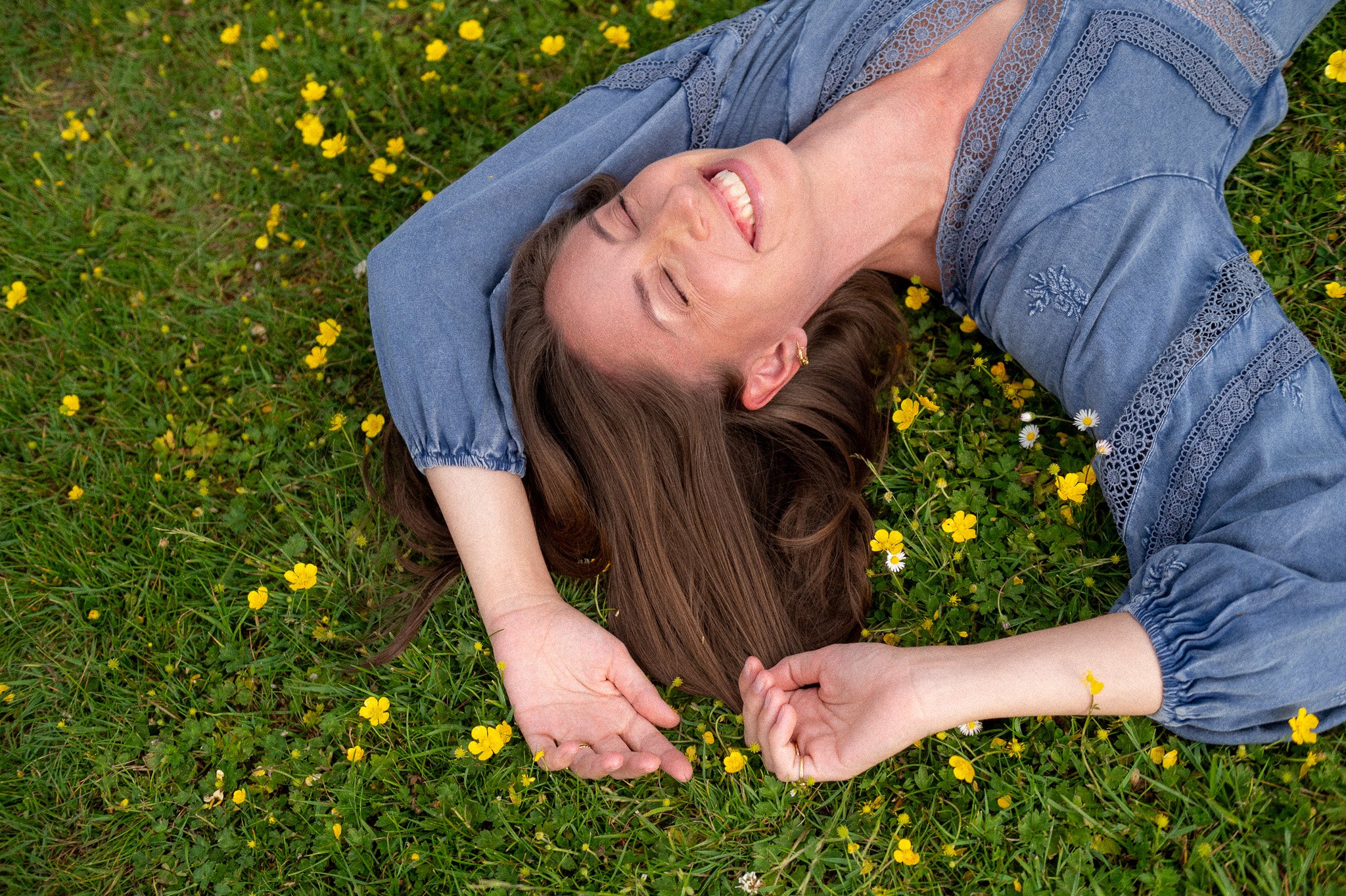A woman with long brown hair lying on grass with small yellow flowers, smiling.