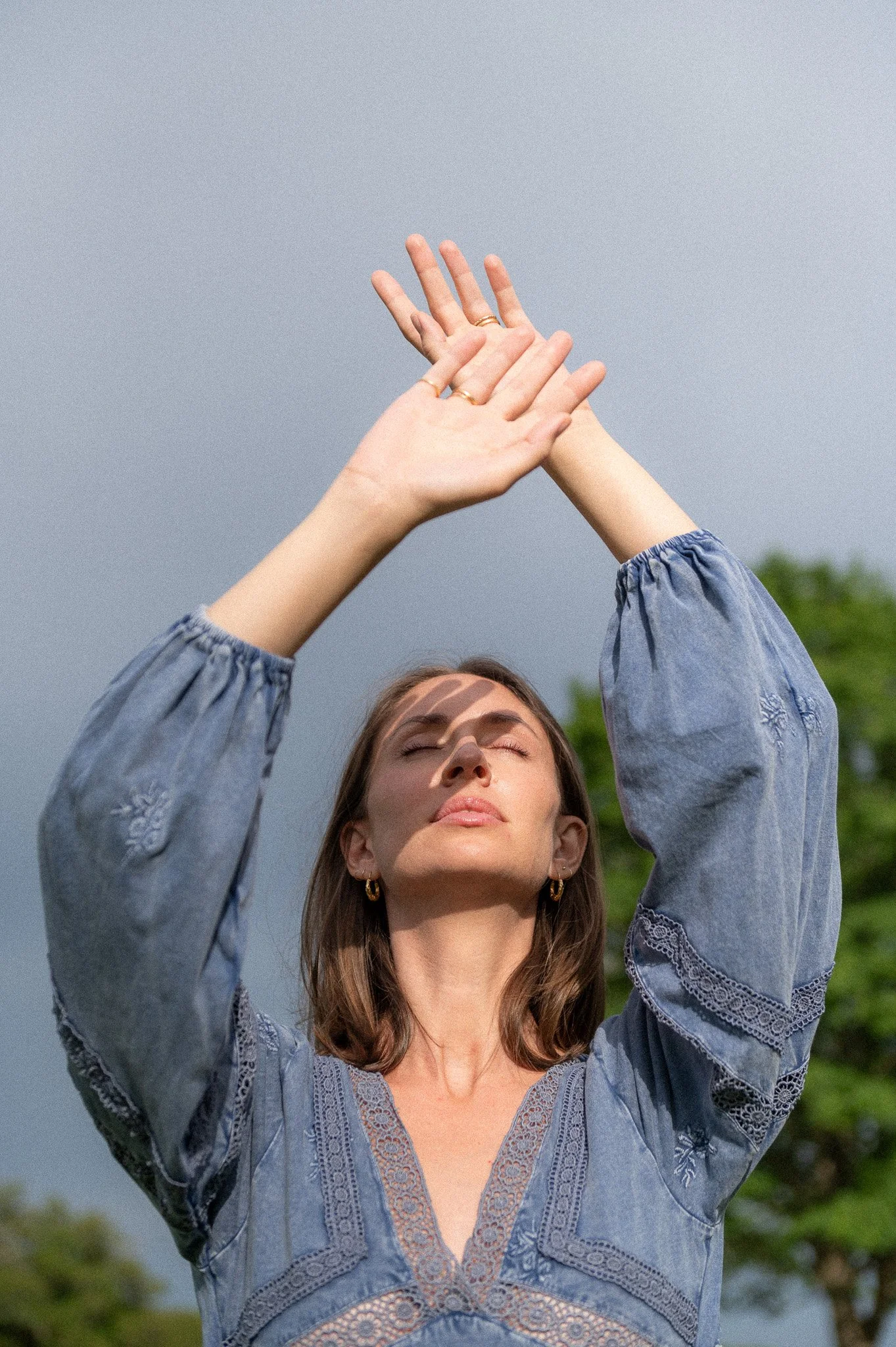 A woman with closed eyes wearing a blue embroidered blouse, holding her hands up in front of a cloudy sky.