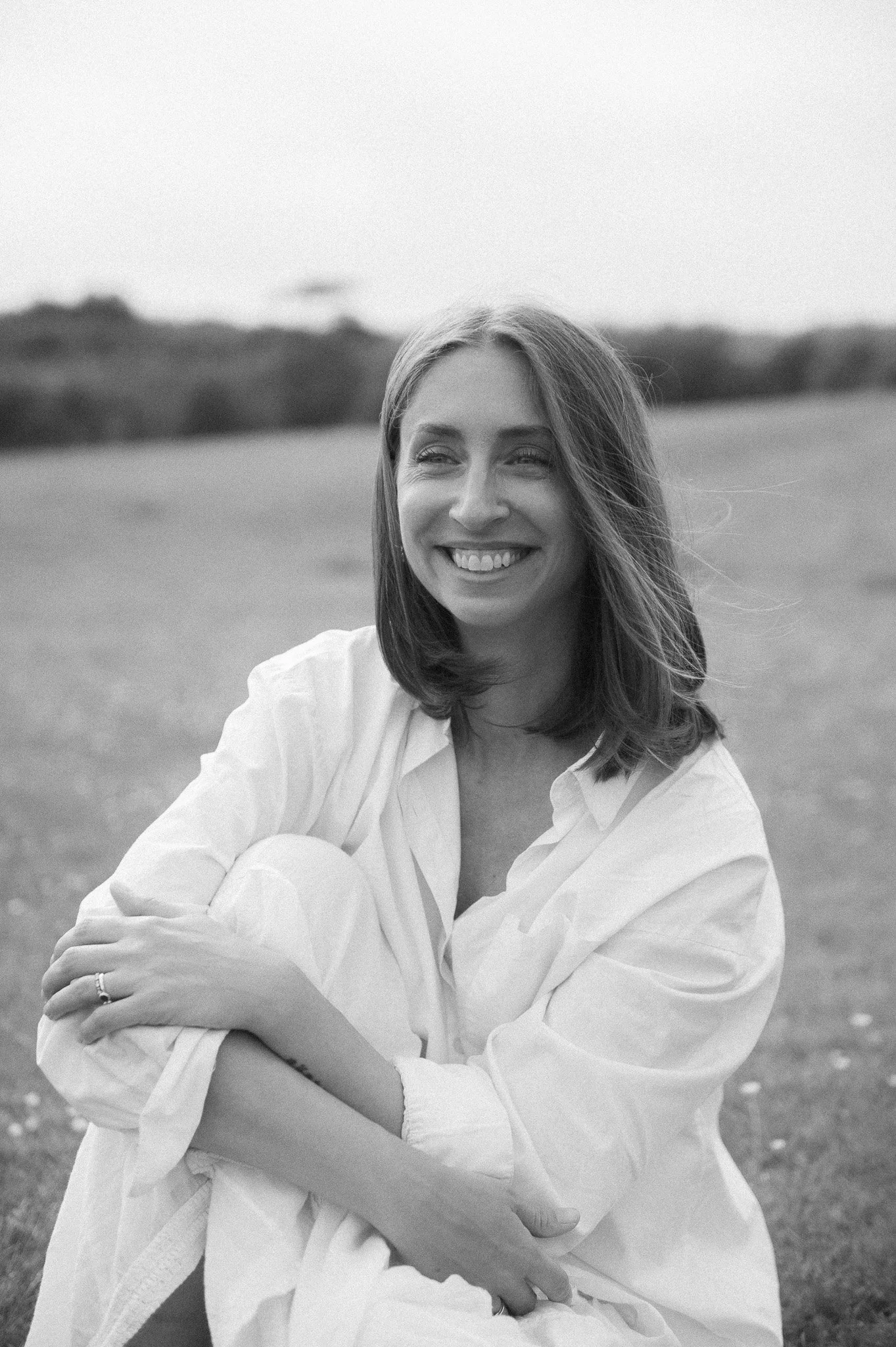 A woman with shoulder-length hair smiling and looking to the side, sitting outdoors in a field.