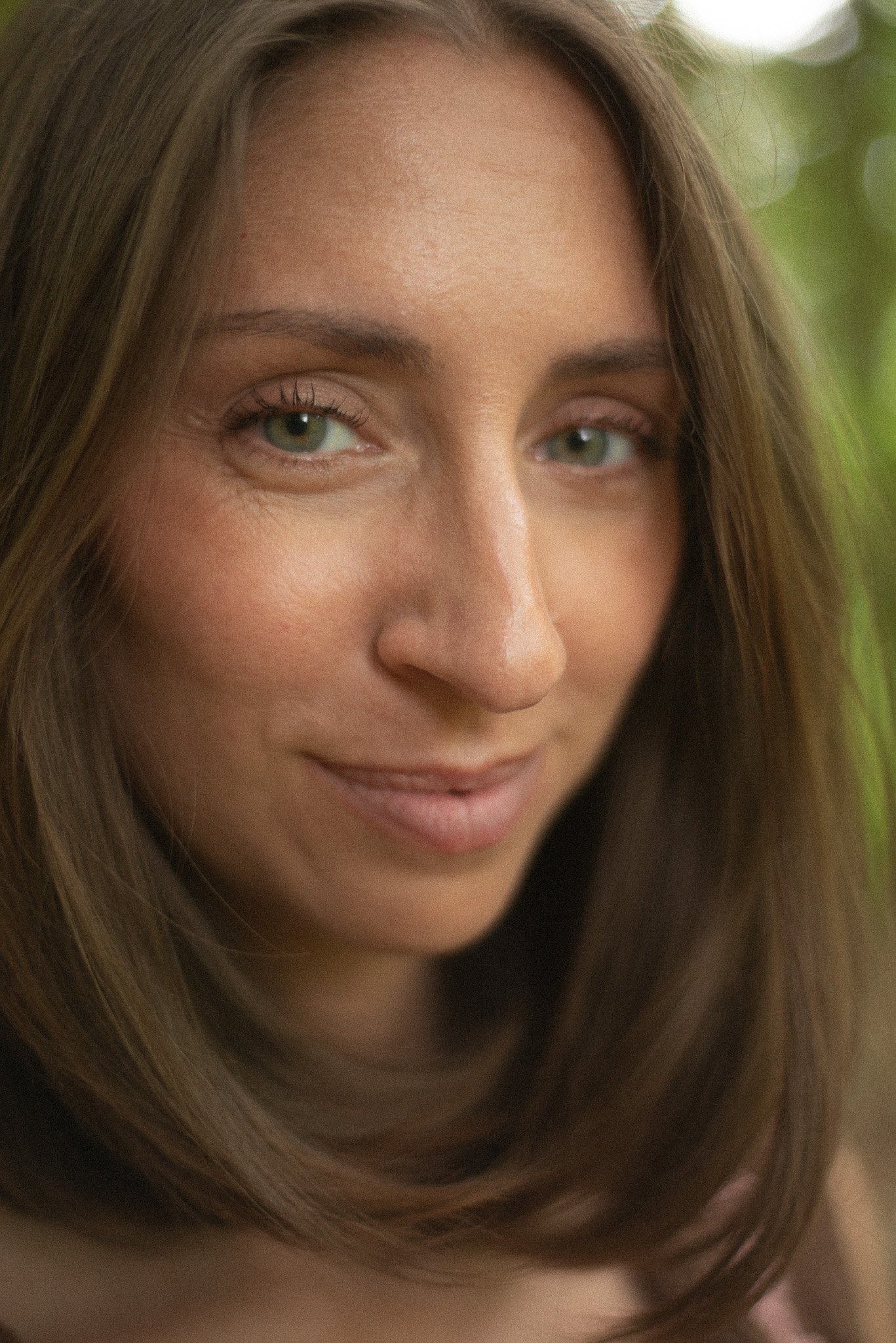 Close-up of a woman's face with light makeup, long brown hair, and light green eyes, smiling softly.