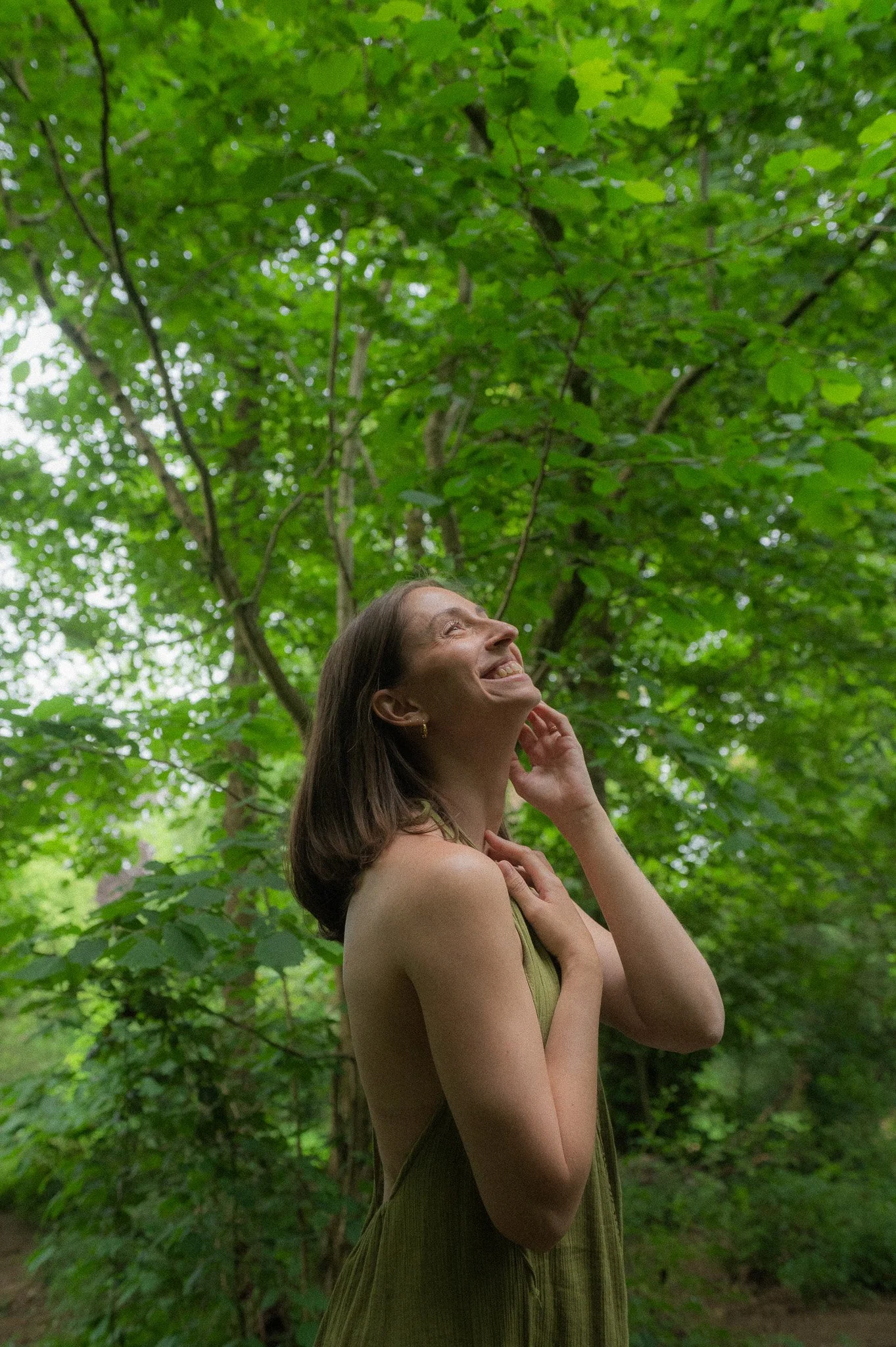 A woman with dark brown, shoulder-length hair looking up and smiling, standing in a lush green forest with trees and leaves in the background.