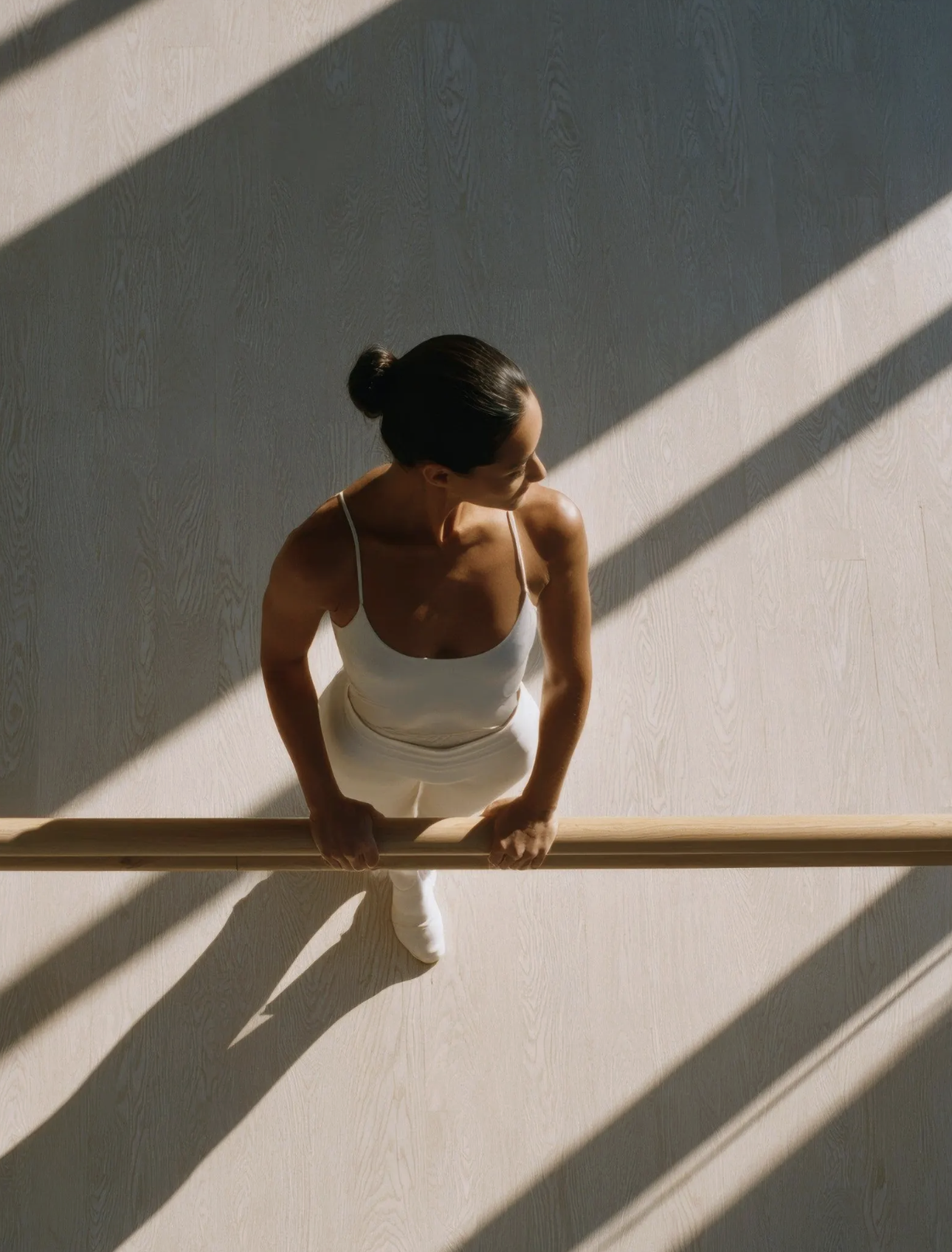 A woman in white workout clothes holding onto a ballet barre in a dance or fitness studio, with sunlight creating diagonal shadows on the light-colored floor and wall.