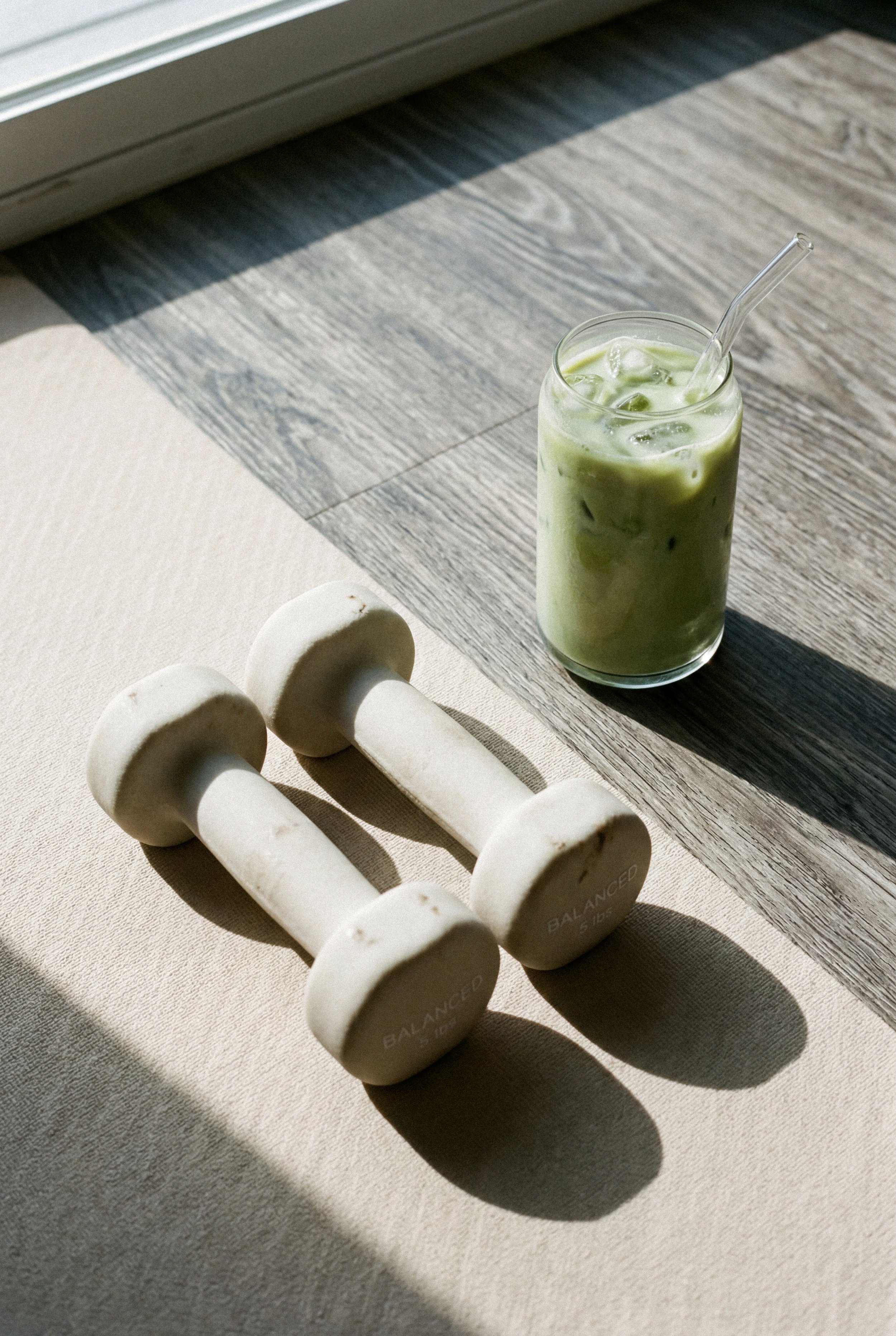 A glass of iced green matcha latte with a straw on a wooden table, next to a white dumbbell with the brand name 'BALANCED' on it, and a white napkin underneath.