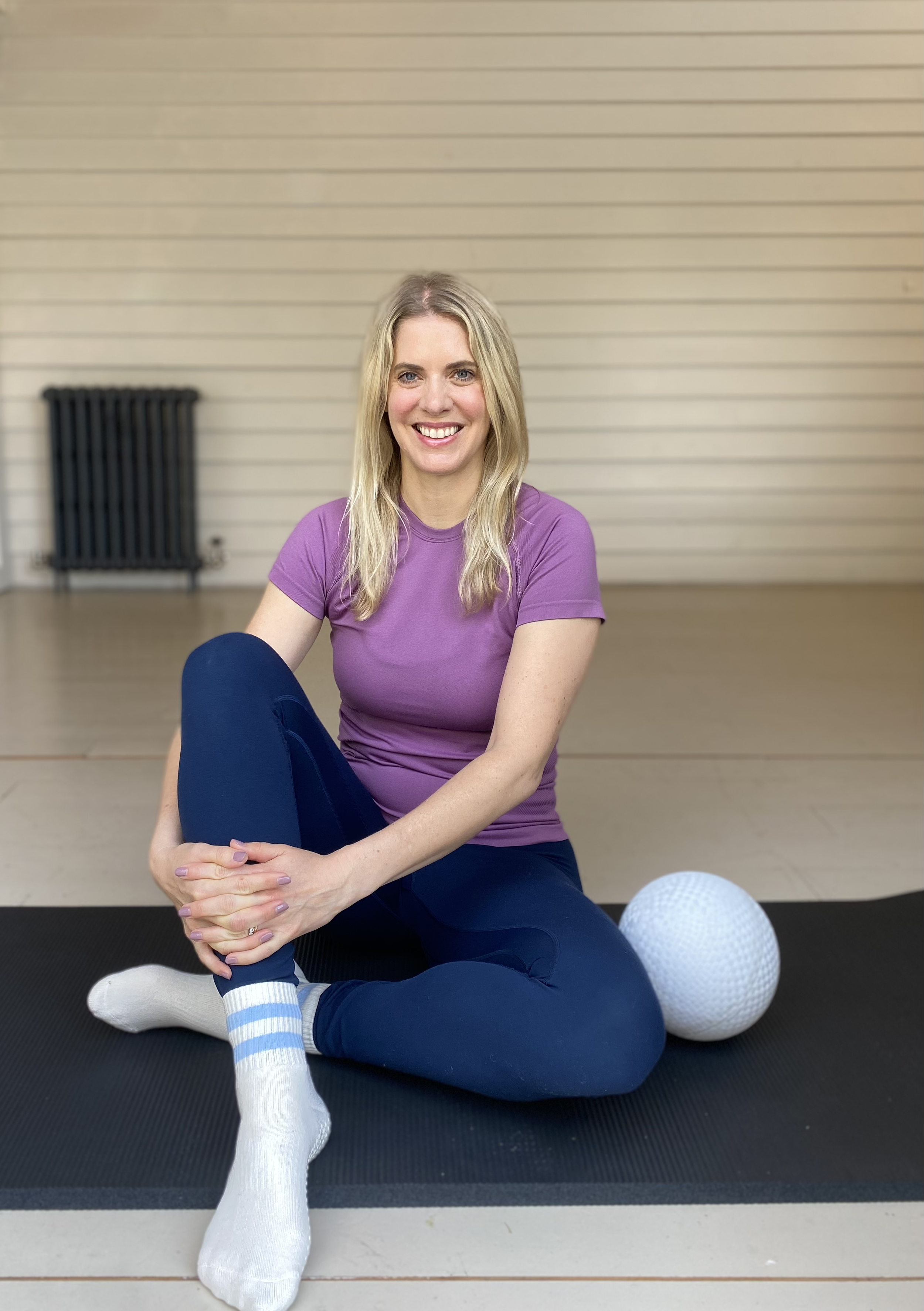 A woman sitting on a yoga mat, smiling, in workout attire, with a white textured massage ball near her foot, in a room with beige horizontal panel walls and a black radiator in the background.