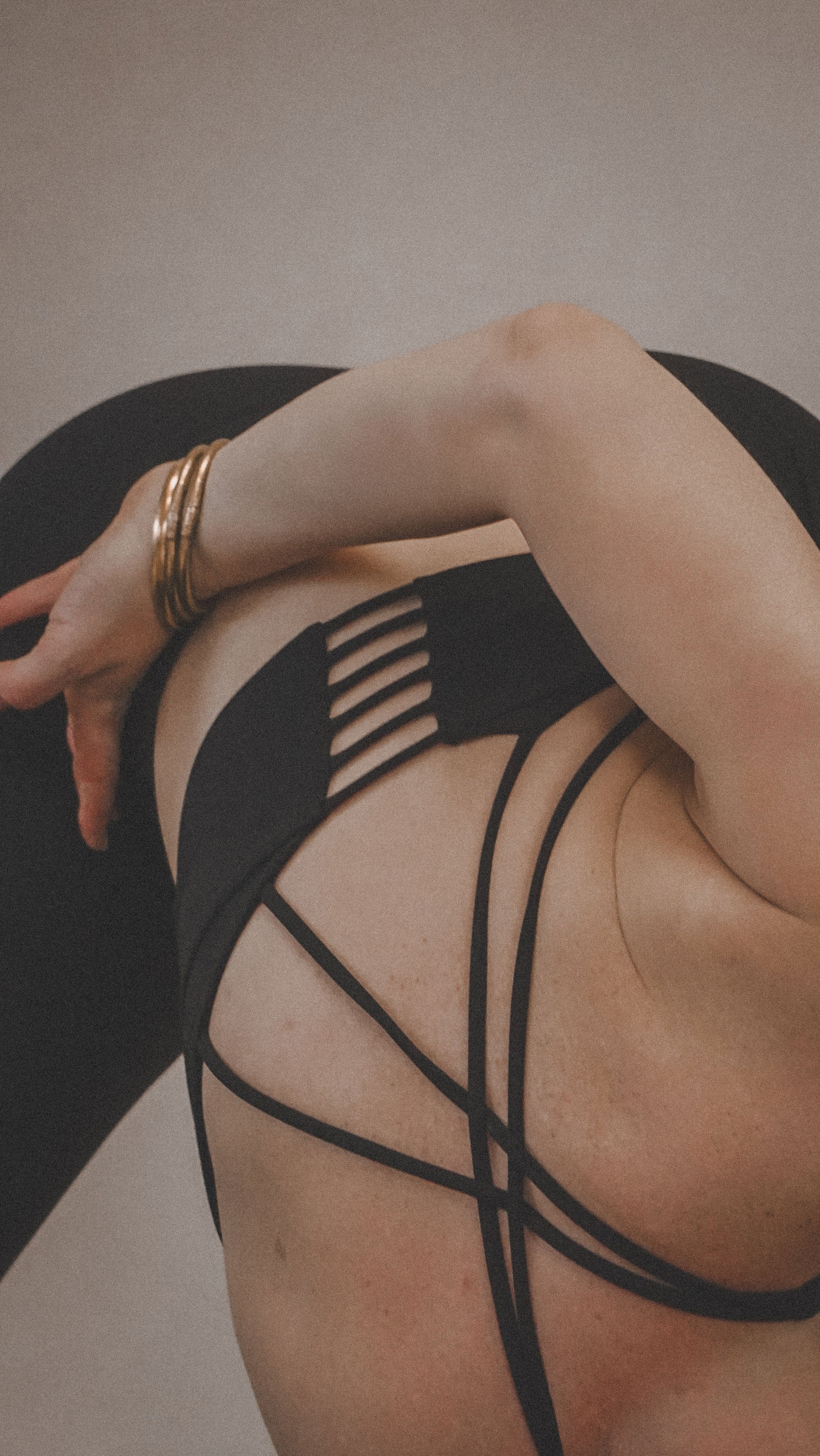 Close-up of a woman reclining on a black chair, wearing black strappy lingerie and gold bangles.