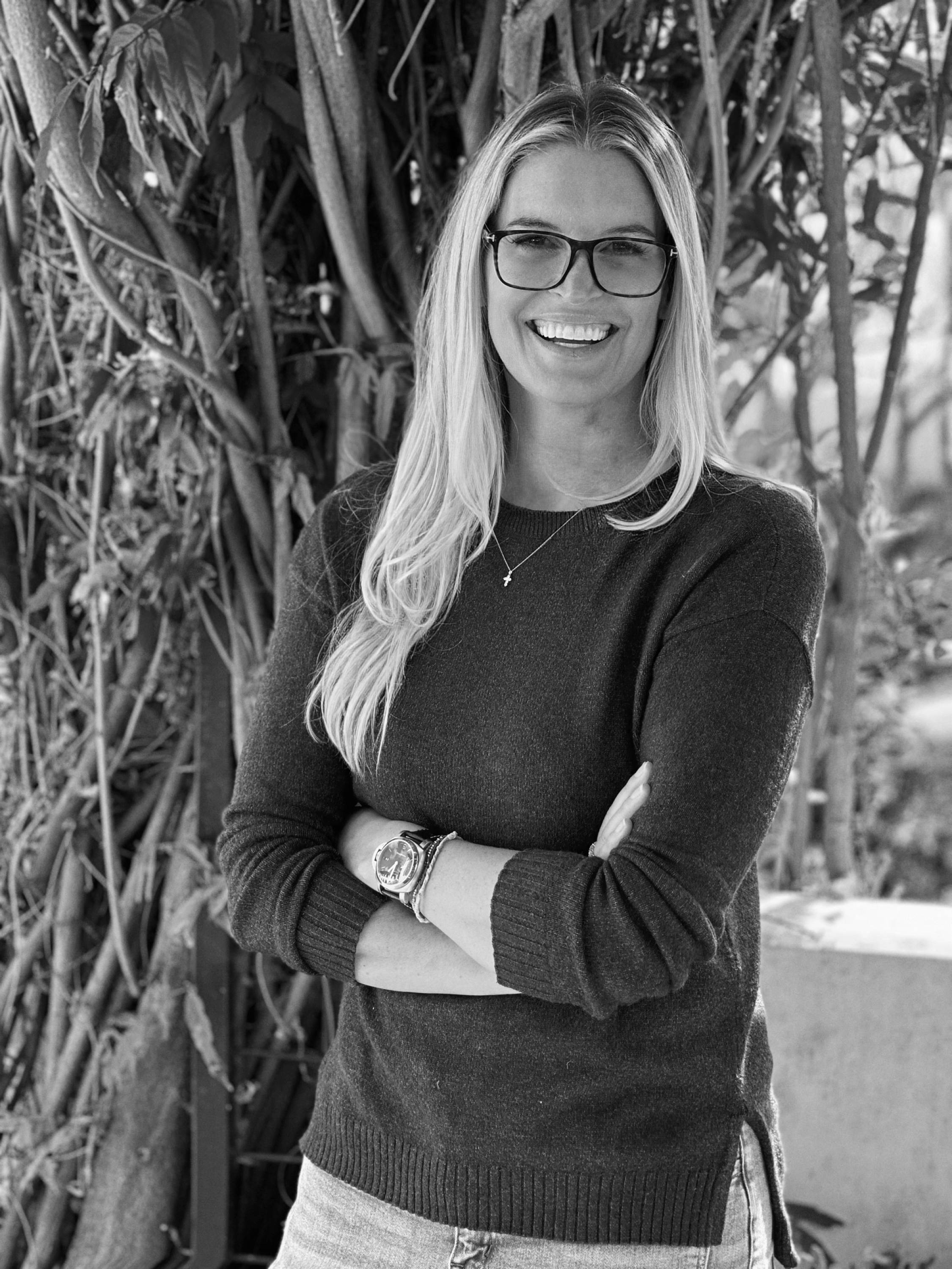 LBUSD School Board Candidate Kimberly Smith smiling, wearing glasses and a dark sweater, standing outdoors with leaves and trees in the background.