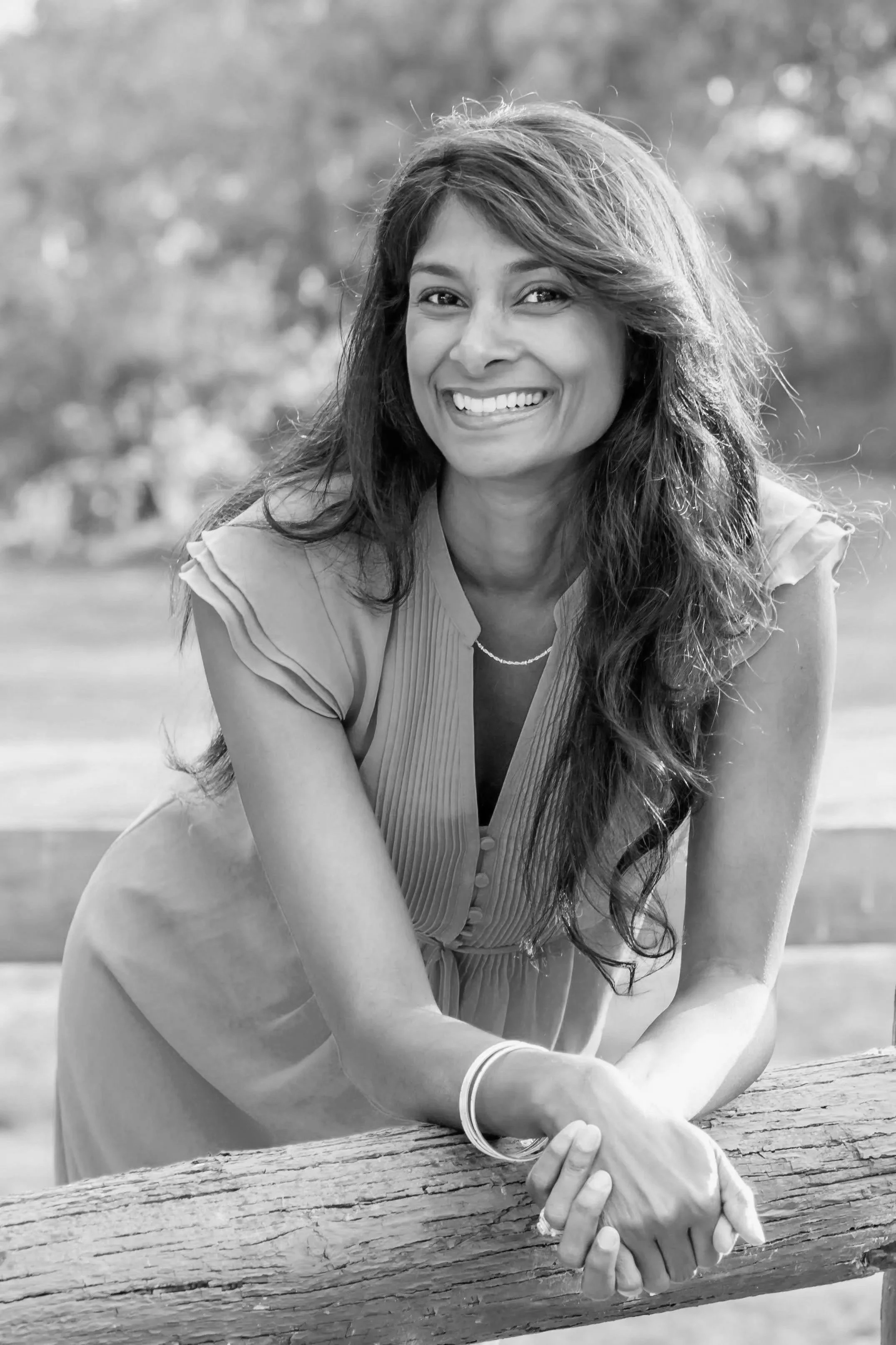 Black and white photo of a LBUSD School Board Candidate Shaheen Sheik-Sadhal with long hair smiling, leaning on a wooden log outdoors.