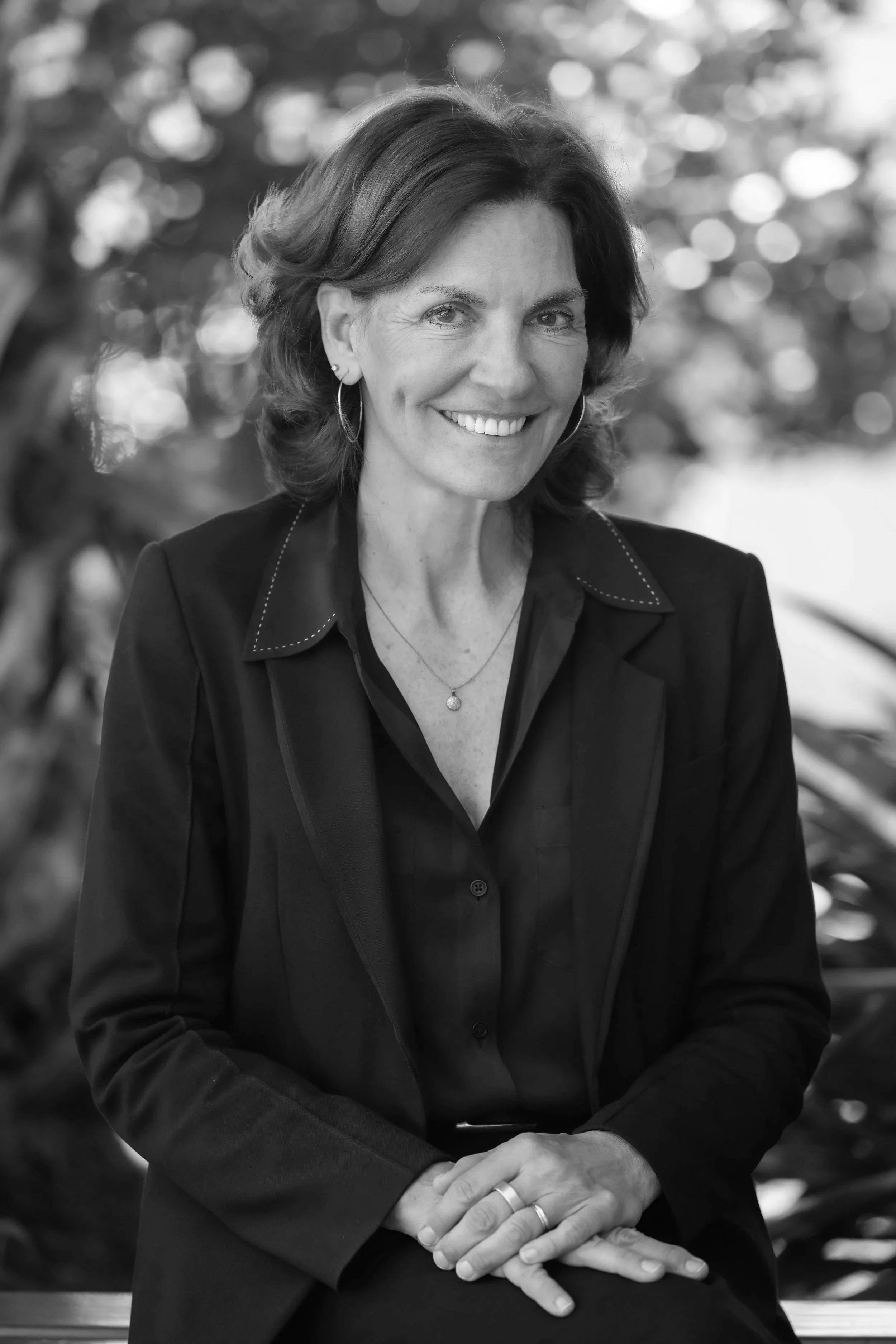 Black and white portrait of a smiling woman with short dark hair, wearing earrings, a necklace, and a blazer, sitting outdoors with blurred trees in the background.