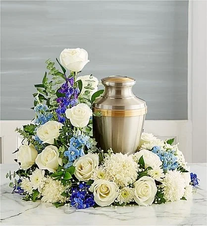 Grayscale urn surrounded by white roses, blue flowers, and greenery on a white surface.