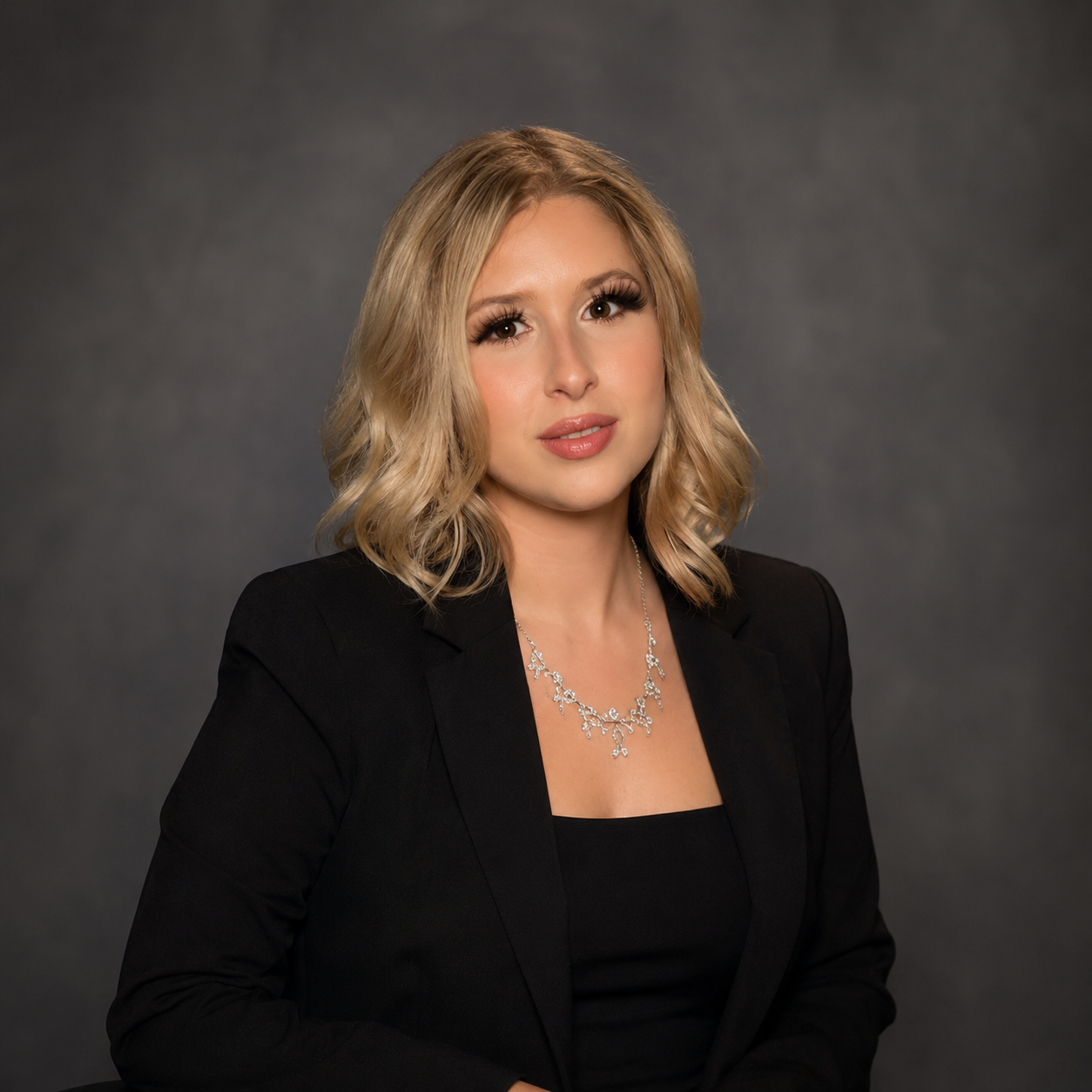 A young woman with blonde hair styled in loose waves, wearing a black blazer and top, and a silver necklace with intricate designs, posing against a dark grey background.