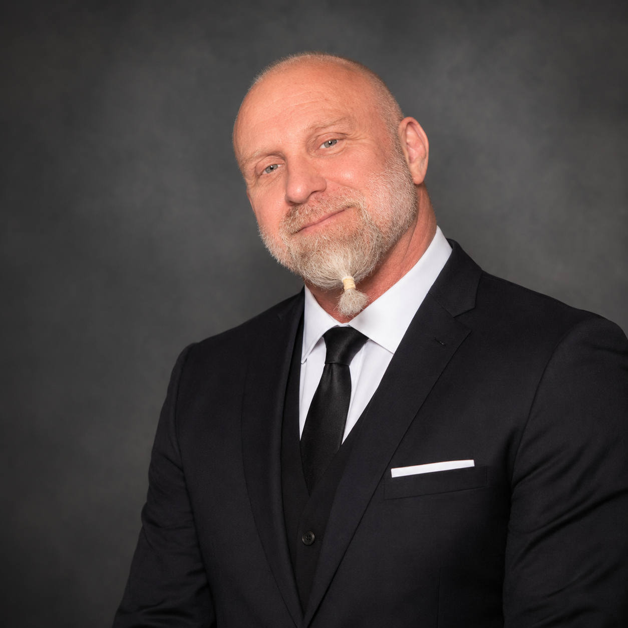Co-owner, Ryan Lykins in a formal black suit, white shirt, and black tie posing against a dark grey background.