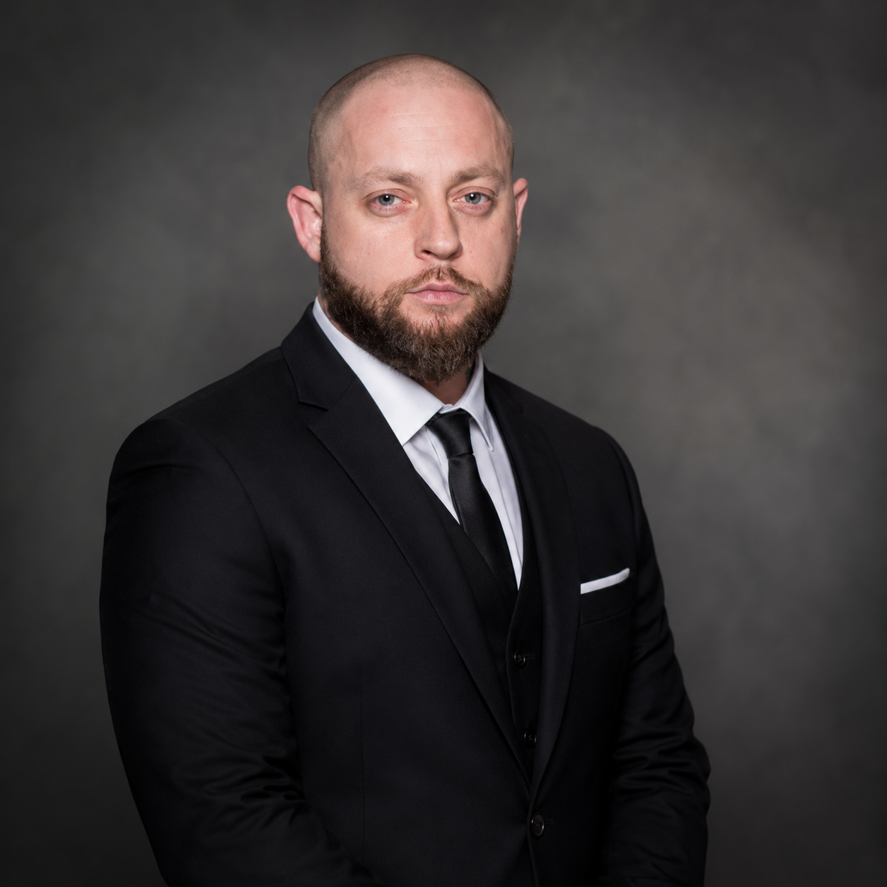 Co-owner, Phil Scheibe wearing a black suit, white shirt, and black tie, standing against a dark gray background.